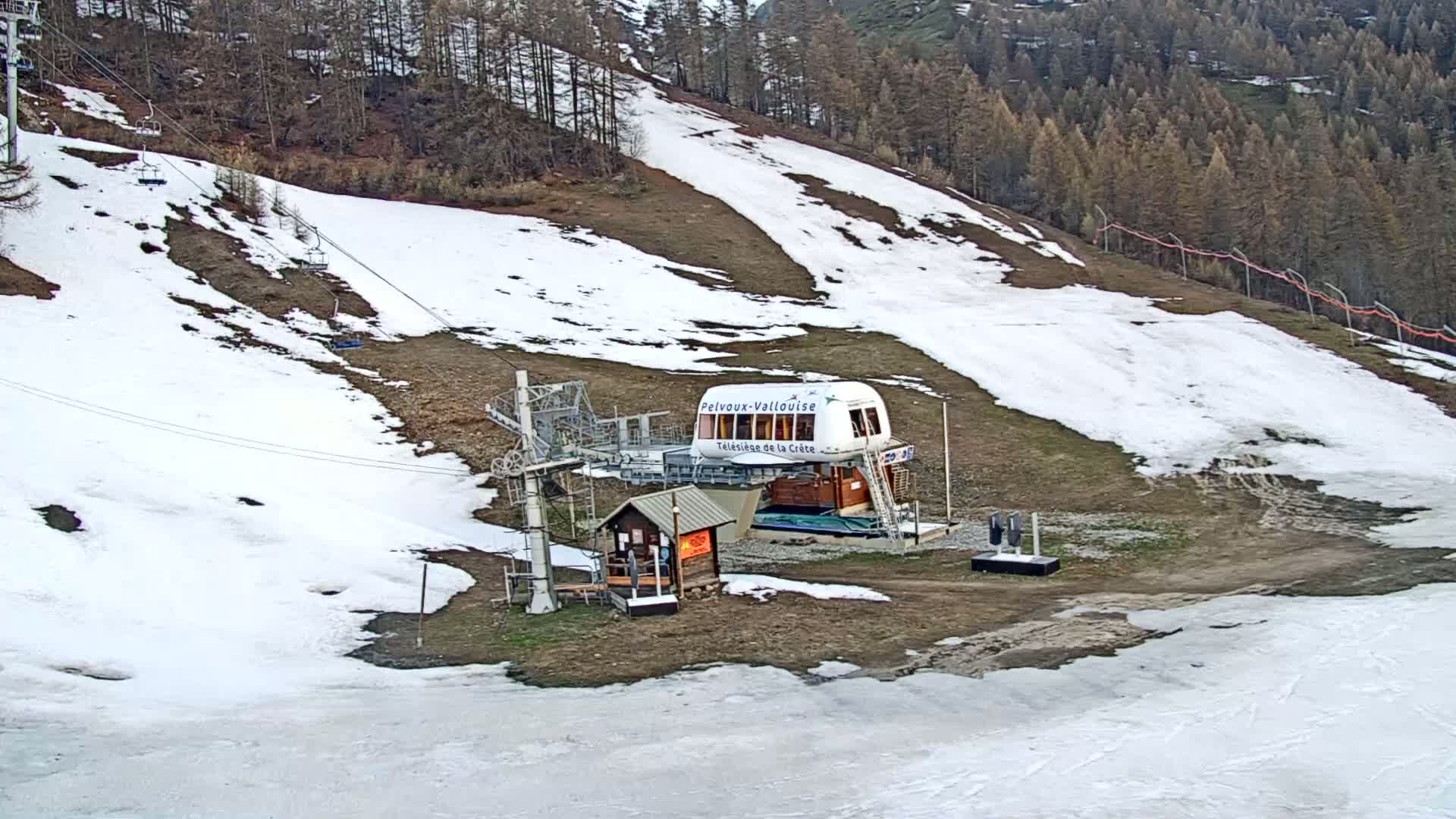 A ski lift station sits on a grassy hillside with patches of snow, under a partly cloudy sky.