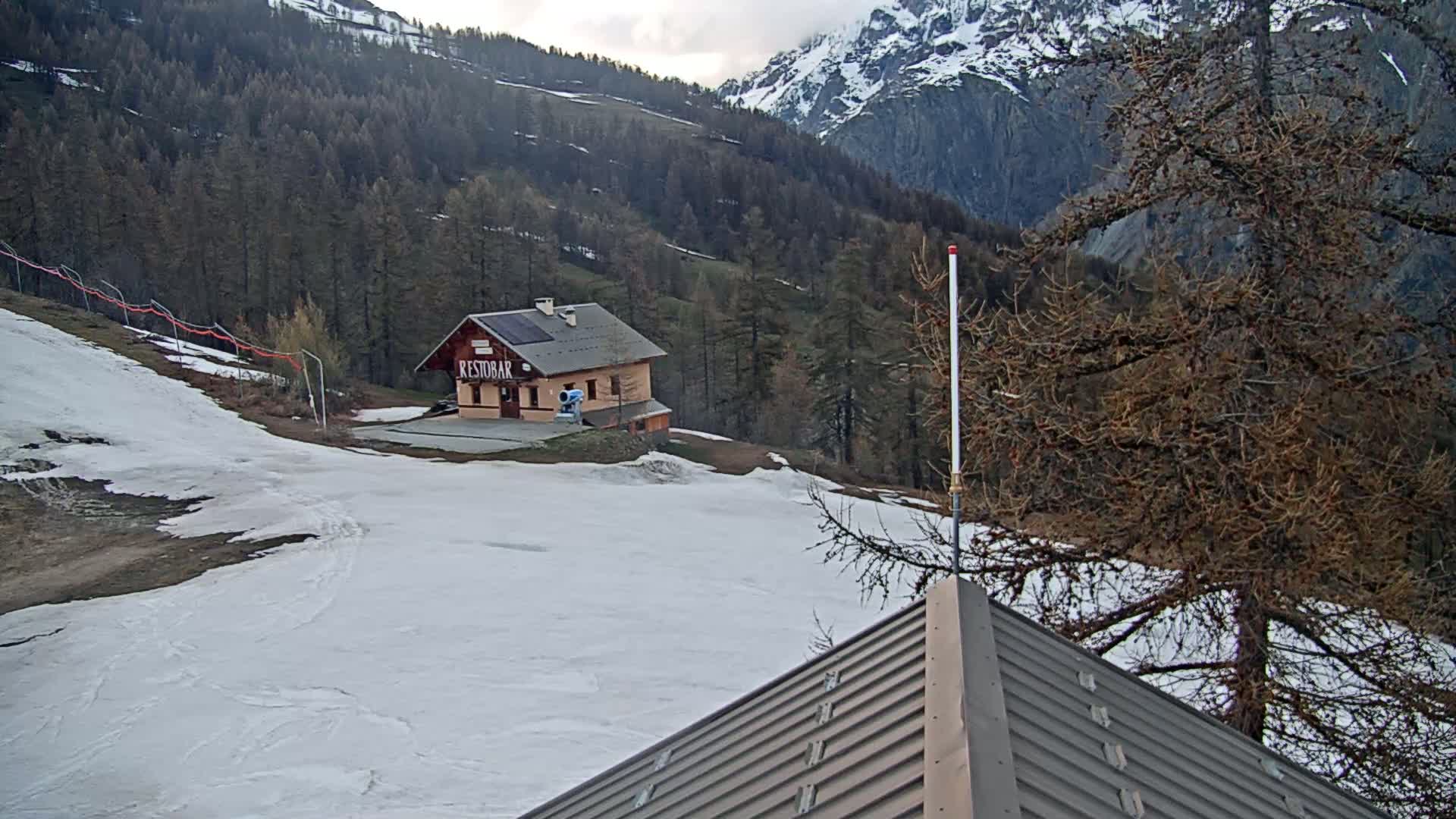 A snow-covered mountain slope with a small building and sparse trees under a cloudy sky.