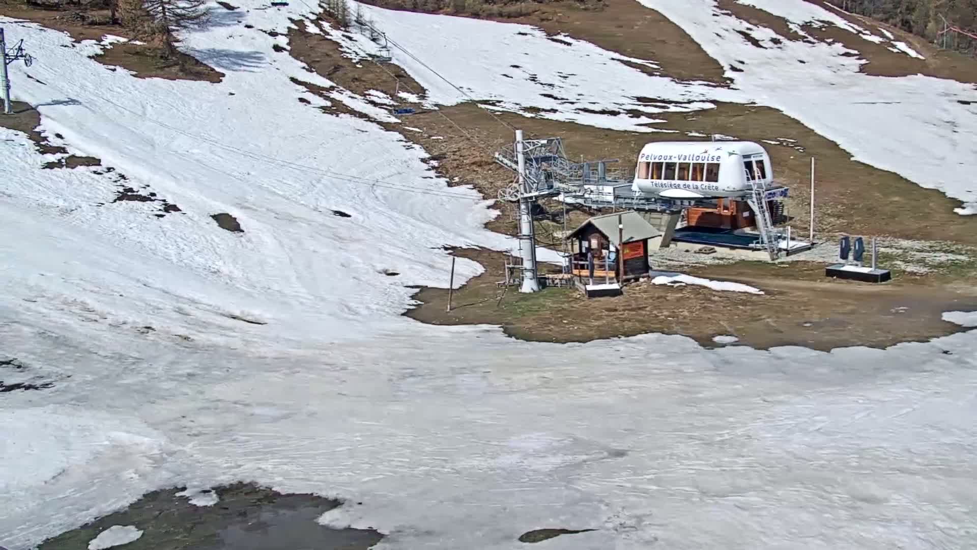 A ski lift station with a small building sits on a mostly brown hillside partially covered in melting snow.