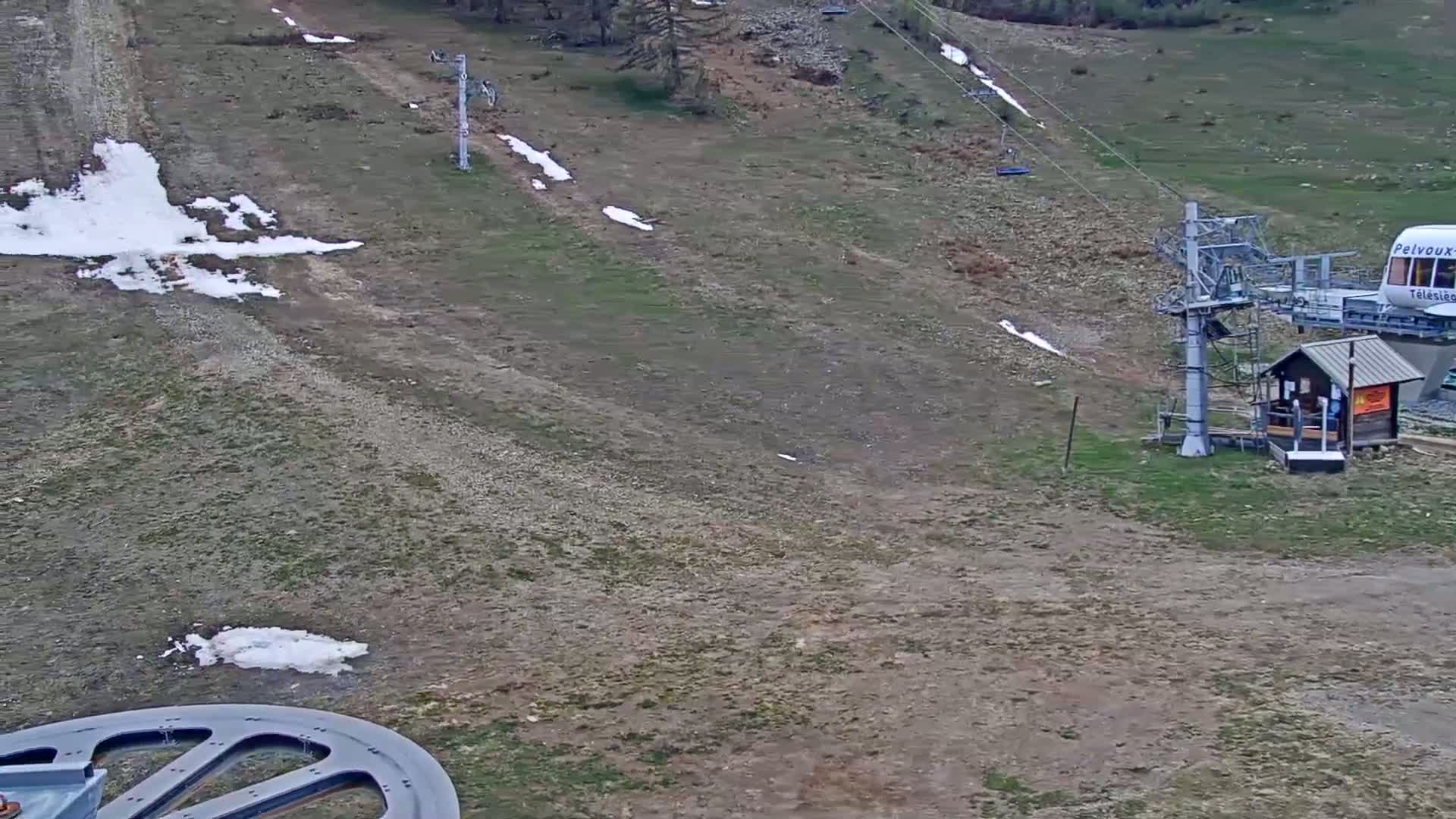 A chairlift and small building sit on a mostly barren hillside with patches of snow and sparse grass, under a seemingly clear sky.