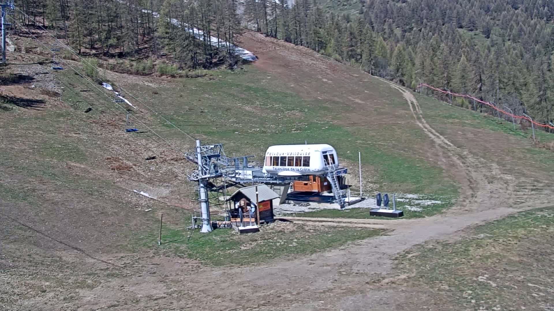 A chairlift station sits on a grassy hillside with a dirt road and sparse, dry vegetation under a sunny sky.