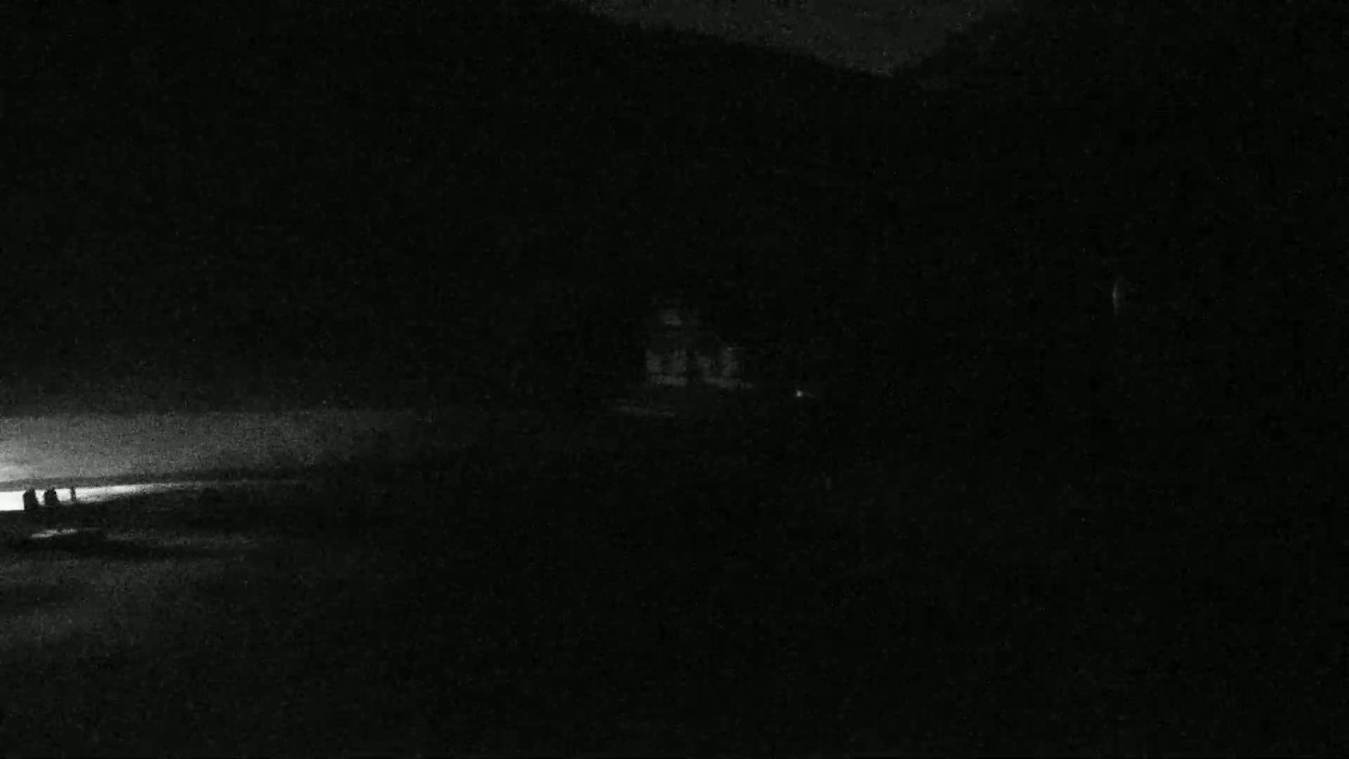 A dimly lit, nighttime scene shows a dark shoreline with a few indistinct figures near a faintly glowing horizon.
