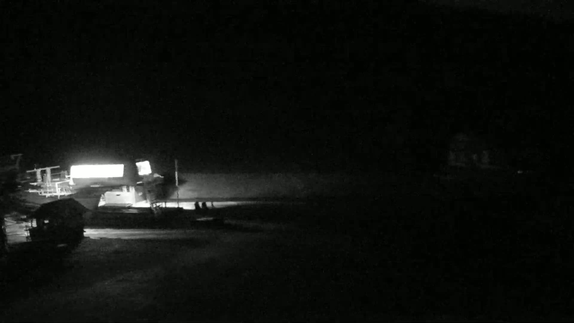 A brightly lit structure and several dark shapes are visible in the nighttime darkness near a body of water.