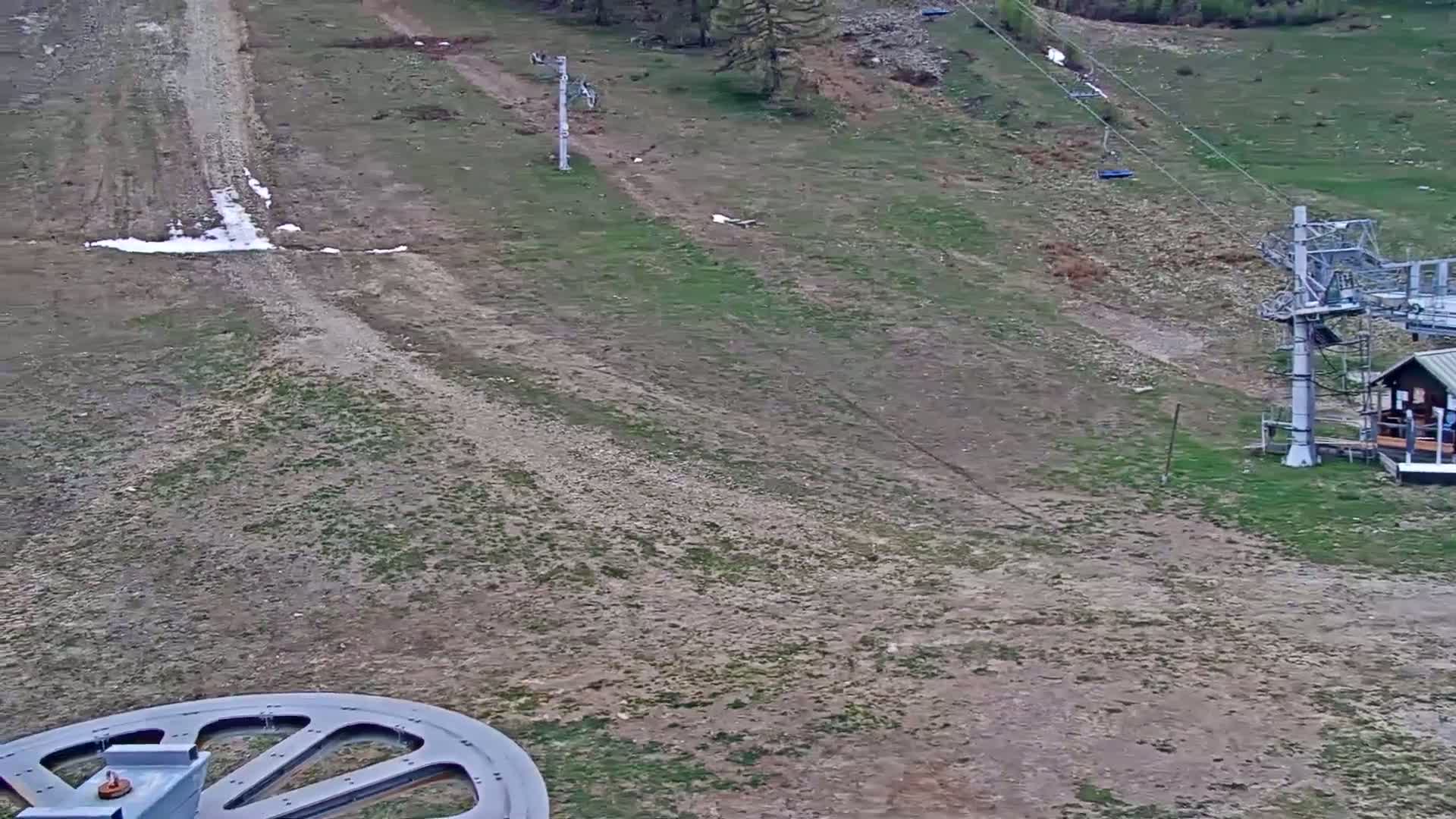 A mostly barren ski slope with patches of grass and some remaining snow, two chairlifts, and a small building are visible in the mostly sunny, spring-like conditions.