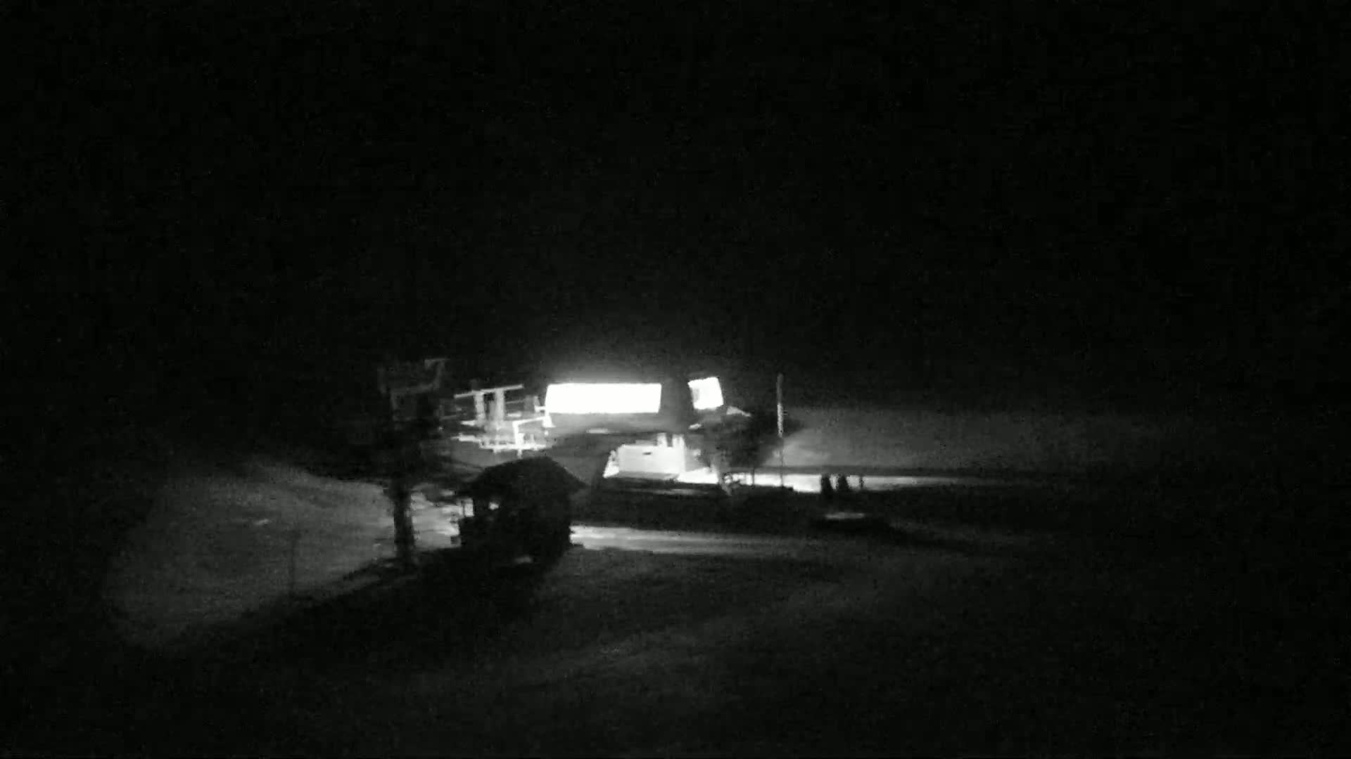 A brightly lit building sits on a snow-covered area at night, with a few shadowy figures and other structures visible nearby.