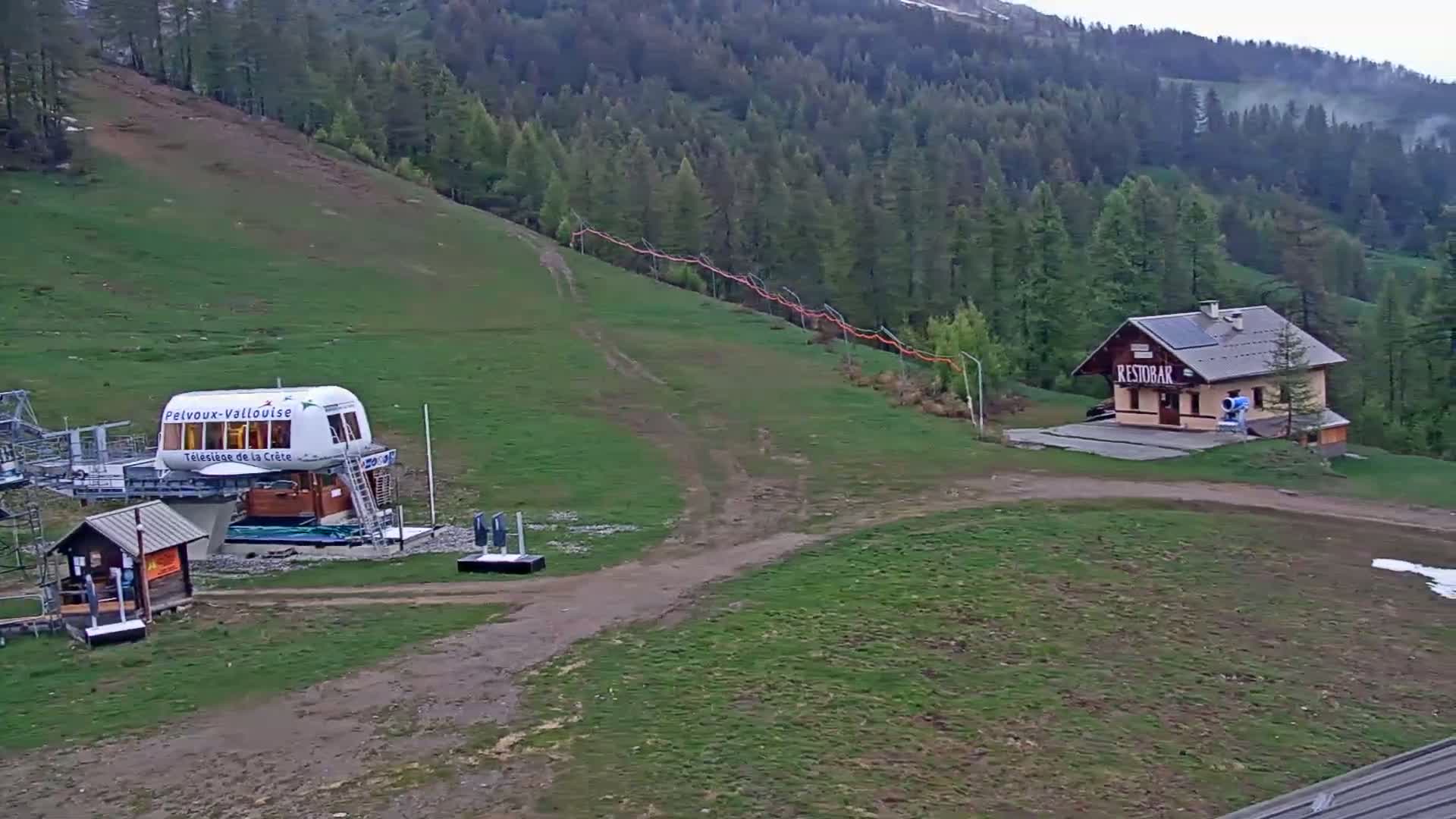 A chairlift station, a small building, and a grassy hill leading up to a coniferous forest under overcast skies.
