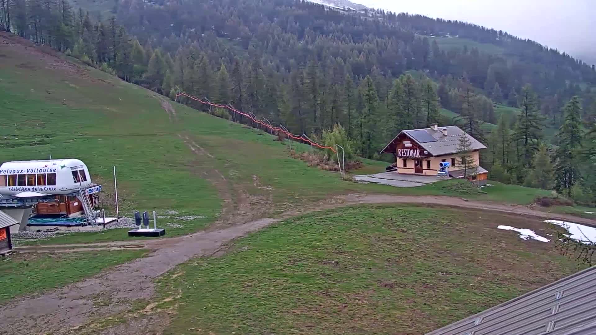 A chairlift base station and small building sit on a grassy hillside overlooking a forested mountain range under an overcast sky.