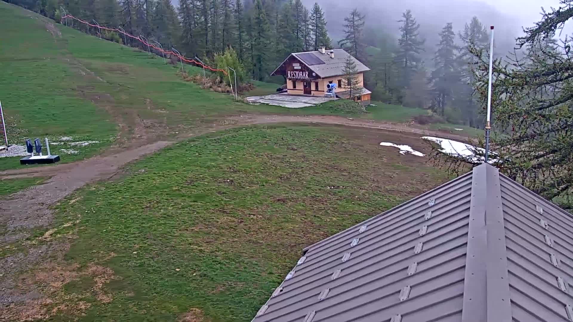 A grassy hillside with a small building, a dirt path, and patches of snow is visible under an overcast sky.