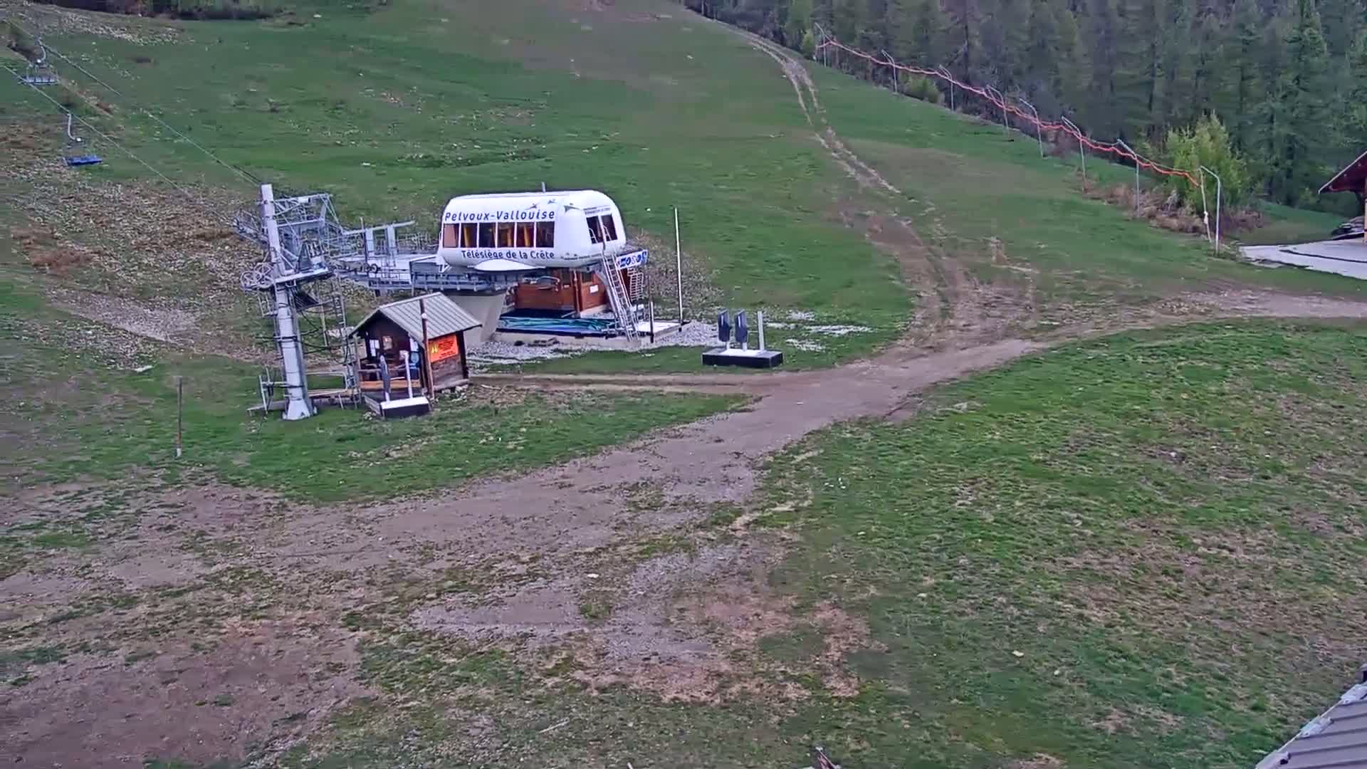 A chairlift station sits on a grassy hillside, with a dirt path leading away from it, under overcast skies.
