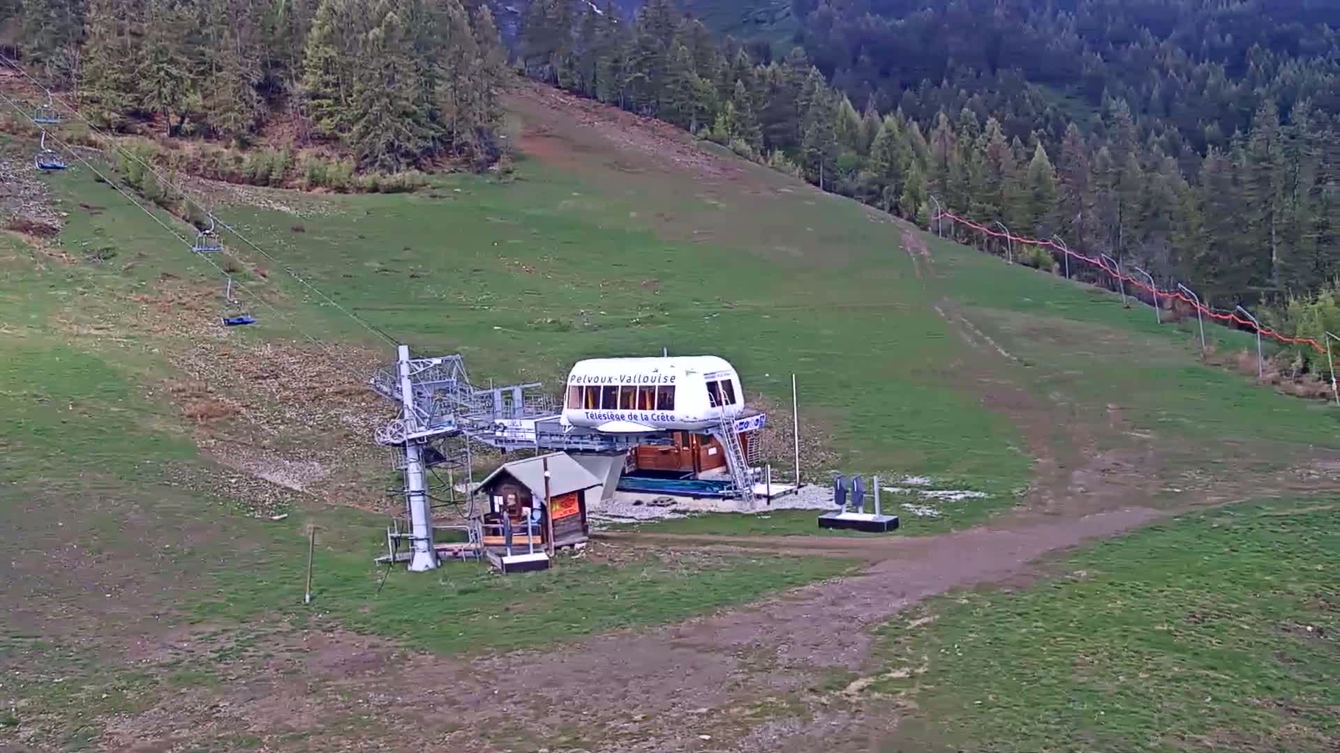 A chairlift station sits on a grassy hillside surrounded by a forest under a partly cloudy sky.