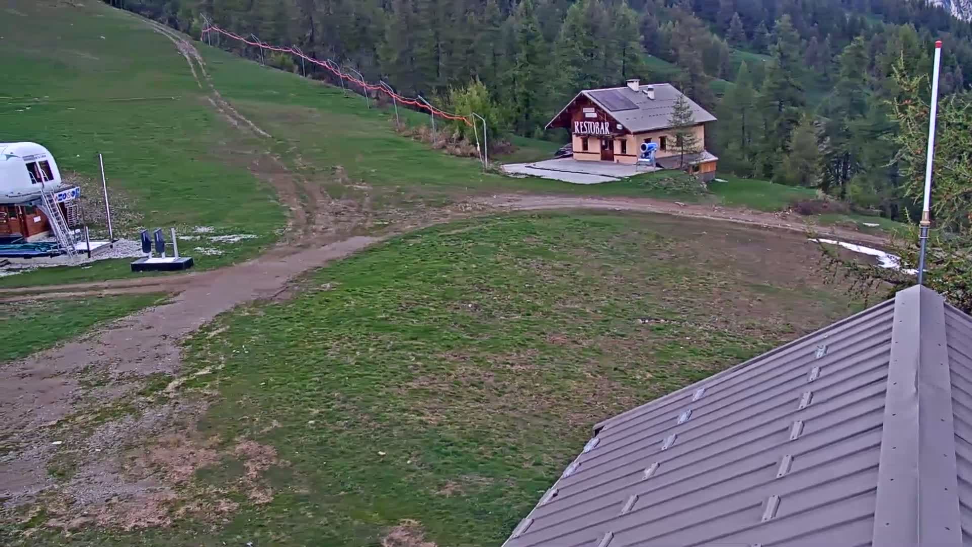 A grassy area with a small building, a dirt path, and a ski lift in the background, under a partly cloudy sky.