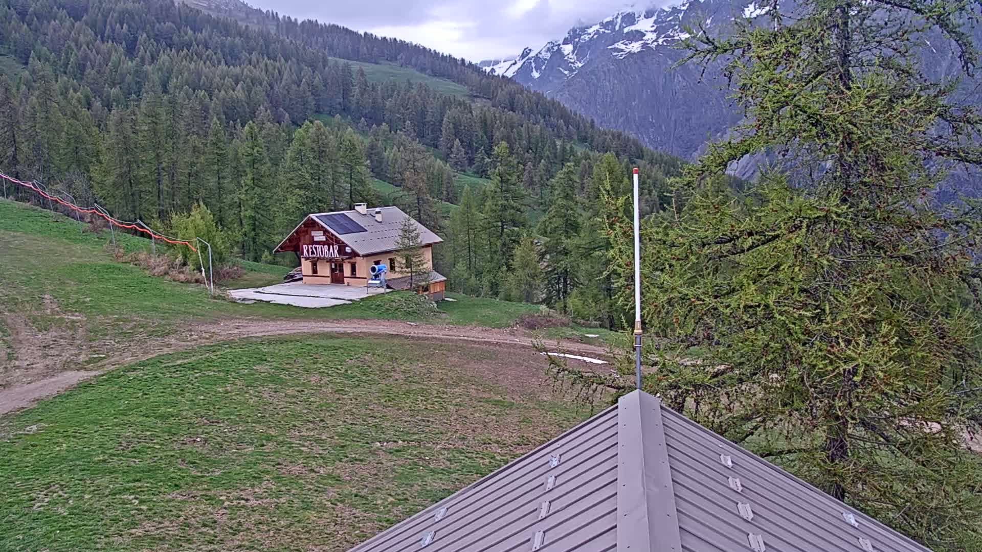 A mountainside view shows a small building among green grass and coniferous trees, with snow-capped mountains in the background under a cloudy sky.