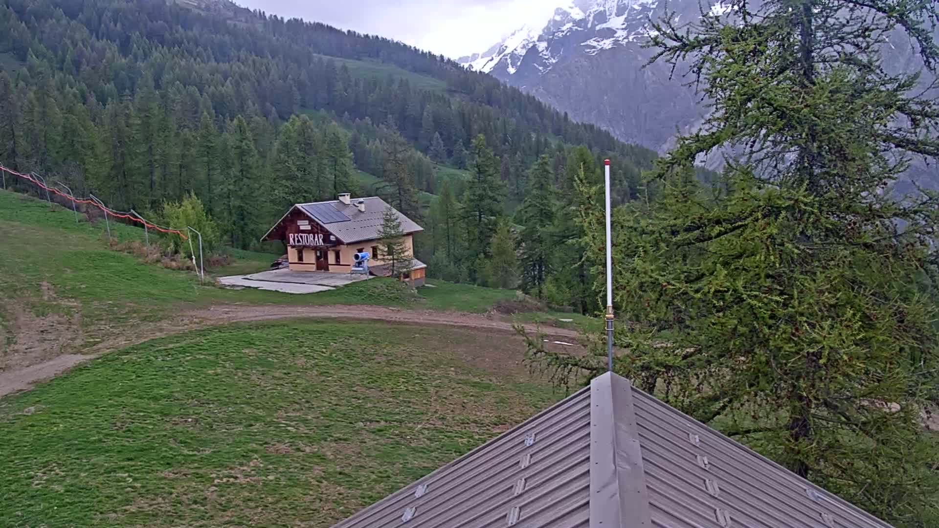 A small building sits on a grassy hillside overlooking a mountain range with snow-capped peaks under an overcast sky.