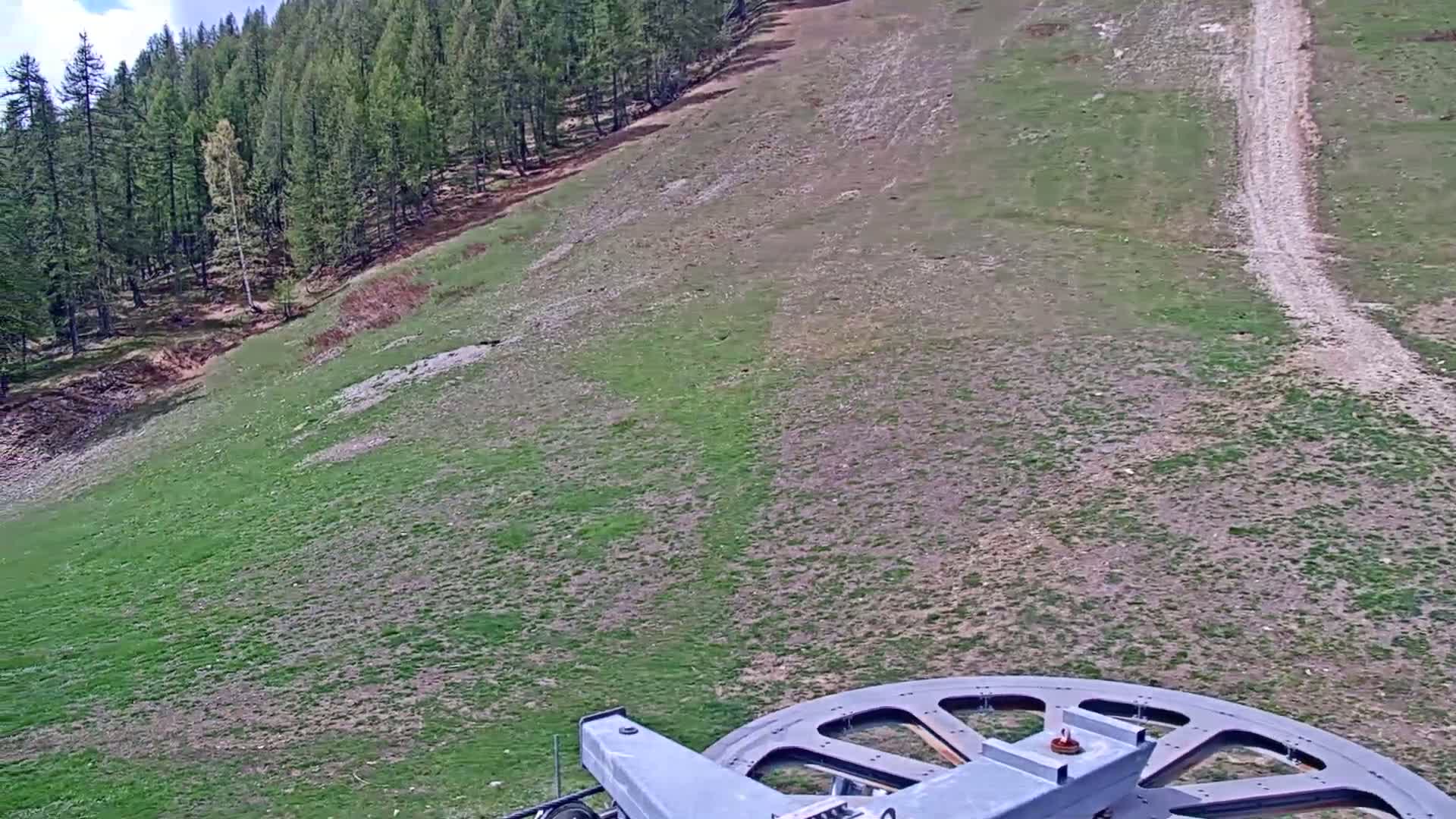 A grassy hillside with a dirt path, partially forested at the top, is viewed from a chairlift.