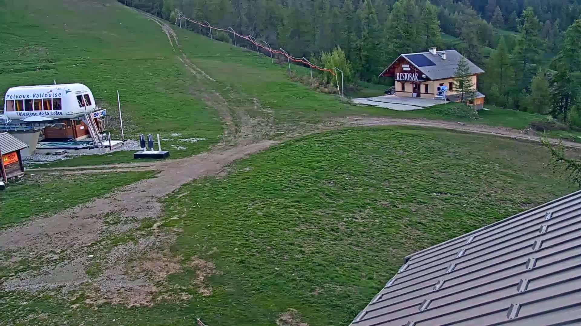 A grassy hillside with a chairlift station, a small building, and a dirt path, under partly sunny skies.