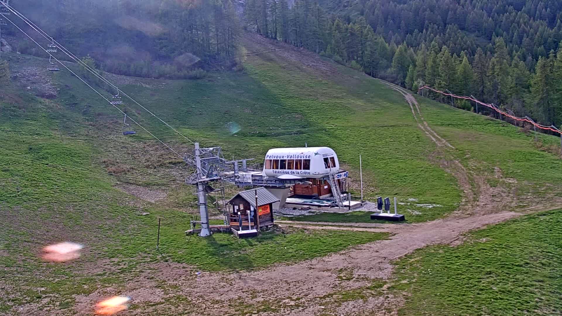 A chairlift station sits on a grassy hillside overlooking a forested mountain, under a mostly sunny sky.
