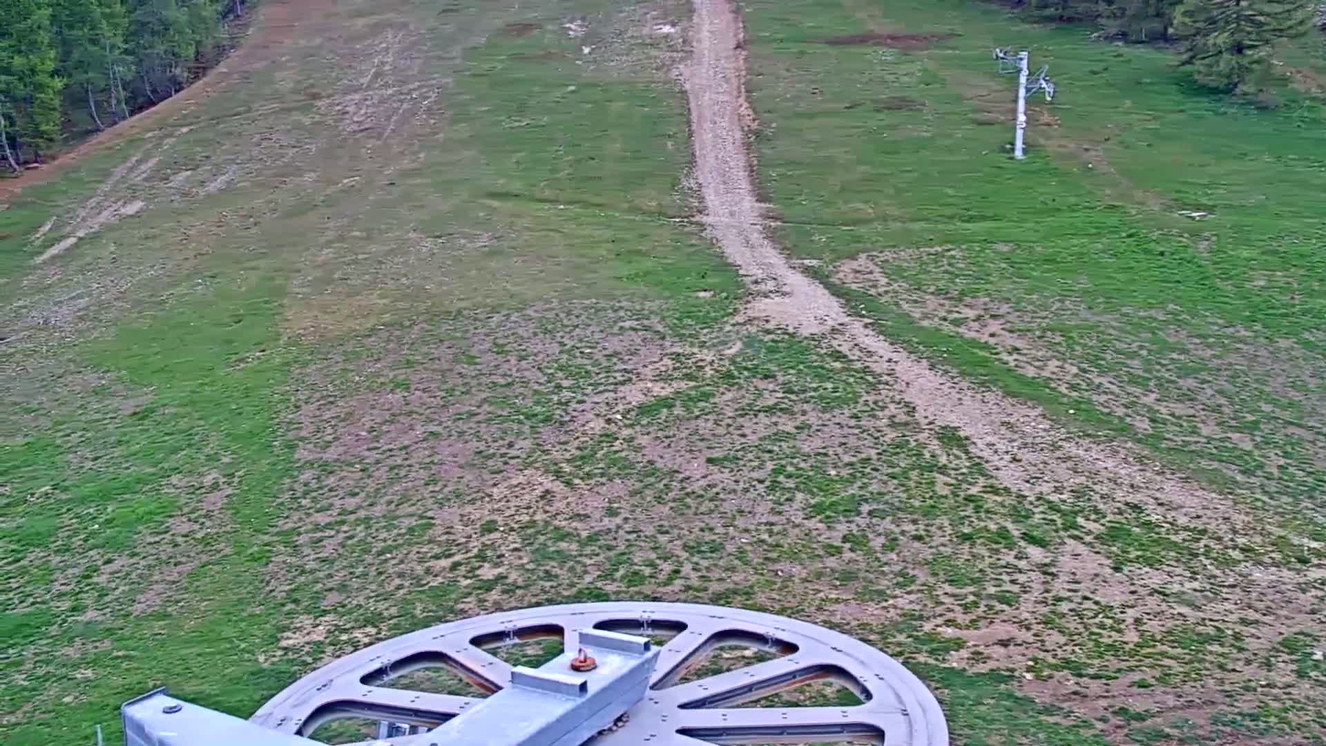 A large wheel of a ski lift is in the foreground overlooking a grassy hill with a dirt path, a ski lift in the distance, and a forest in the background.