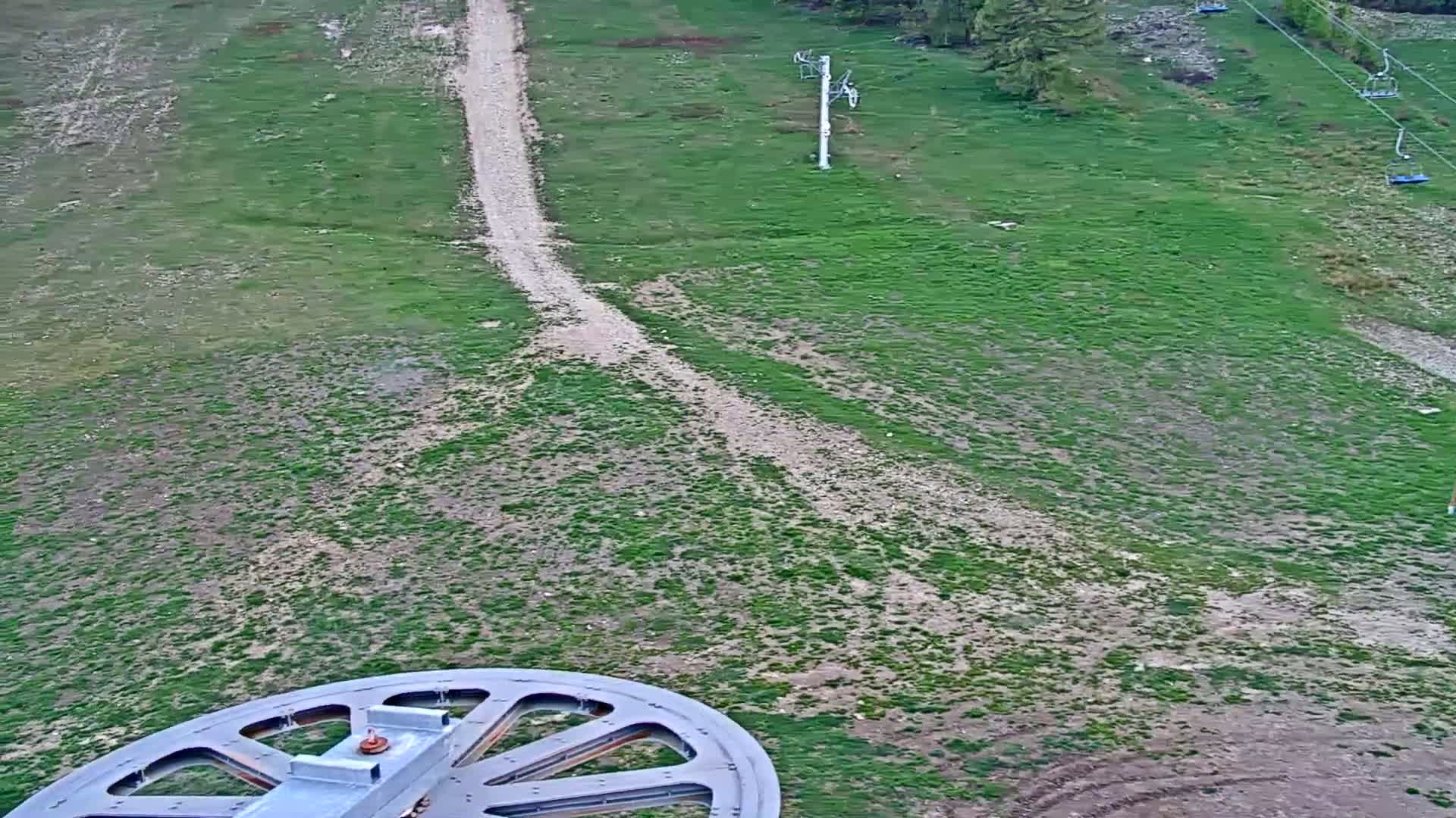 A large wheel is in the foreground, overlooking a grassy hillside with dirt paths and inactive ski lifts.
