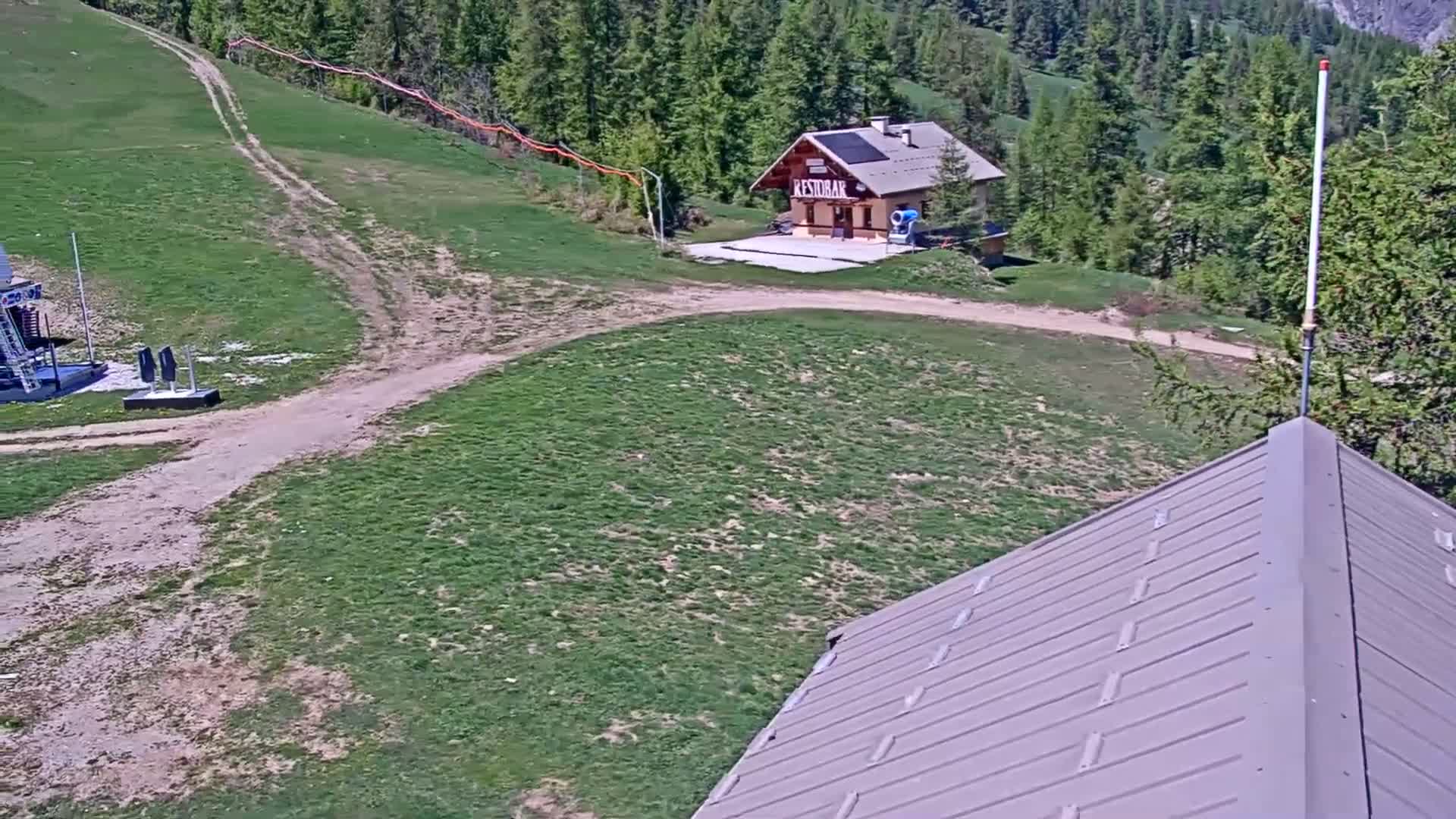 A grassy hillside with a small building, a dirt path, and part of a metal roof are visible on a sunny day.