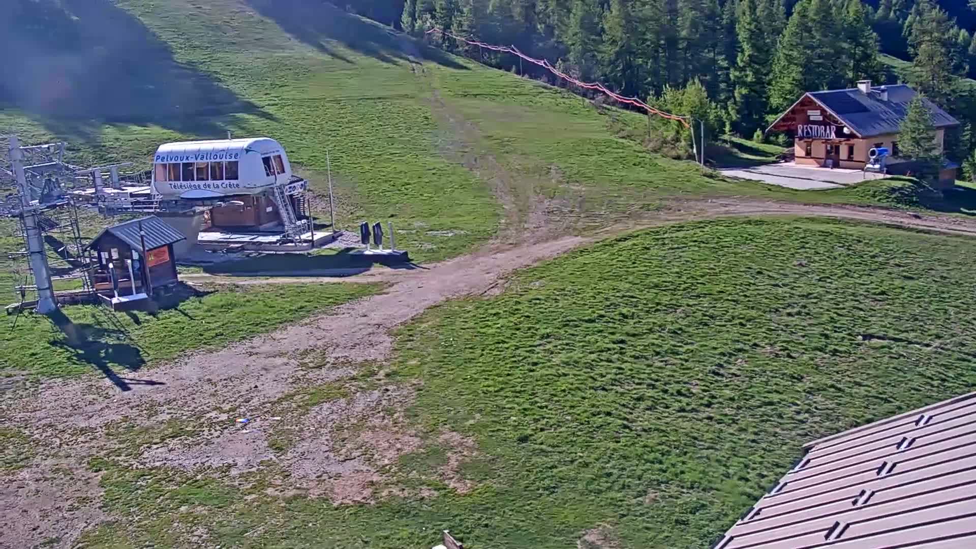 A chairlift station and small building sit on a grassy hillside, with a dirt path leading between them under a sunny sky.
