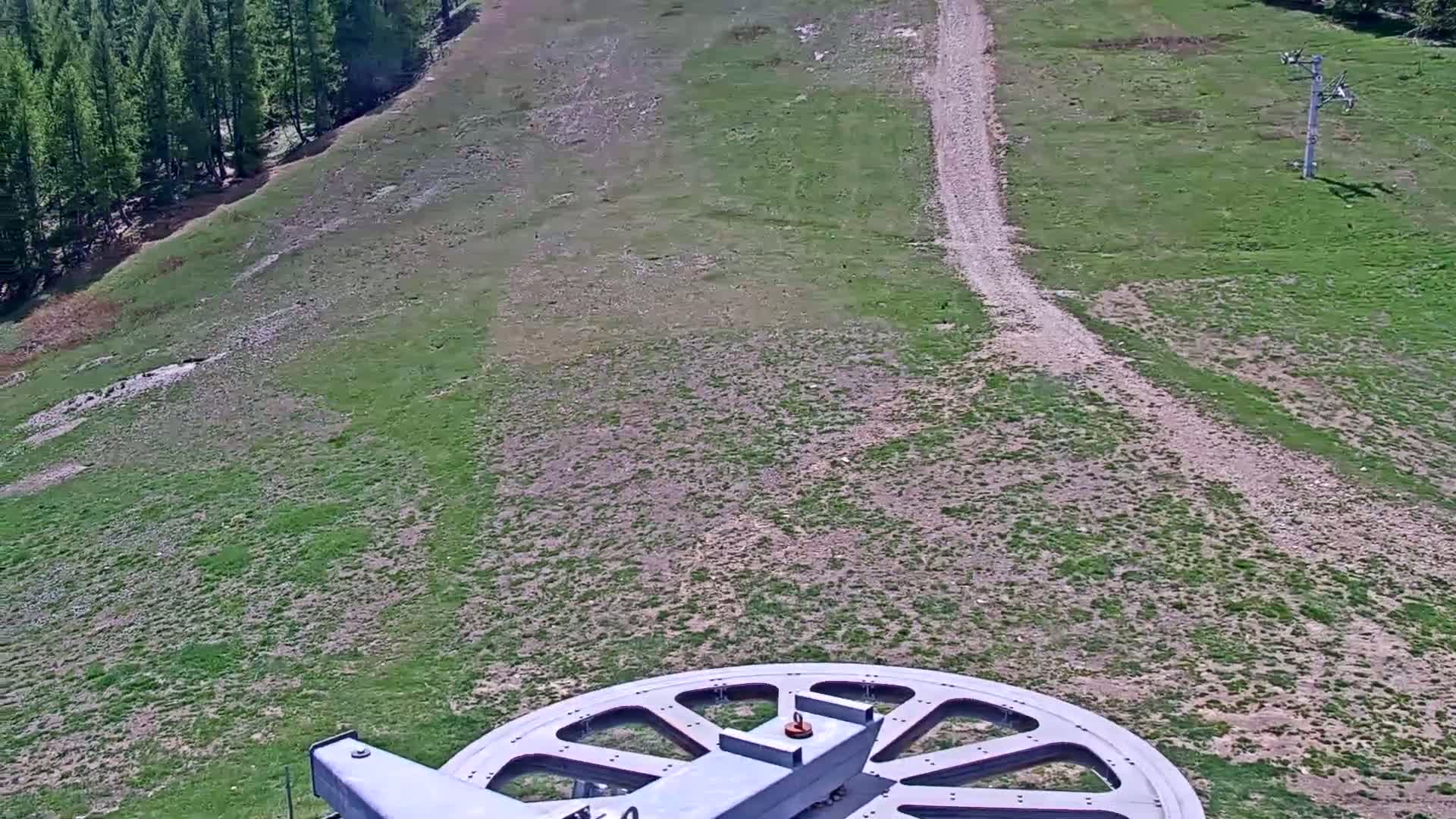 A large metal wheel is partially visible in the foreground, overlooking a grassy hillside with a dirt path and a sparse forest in the distance on a sunny day.