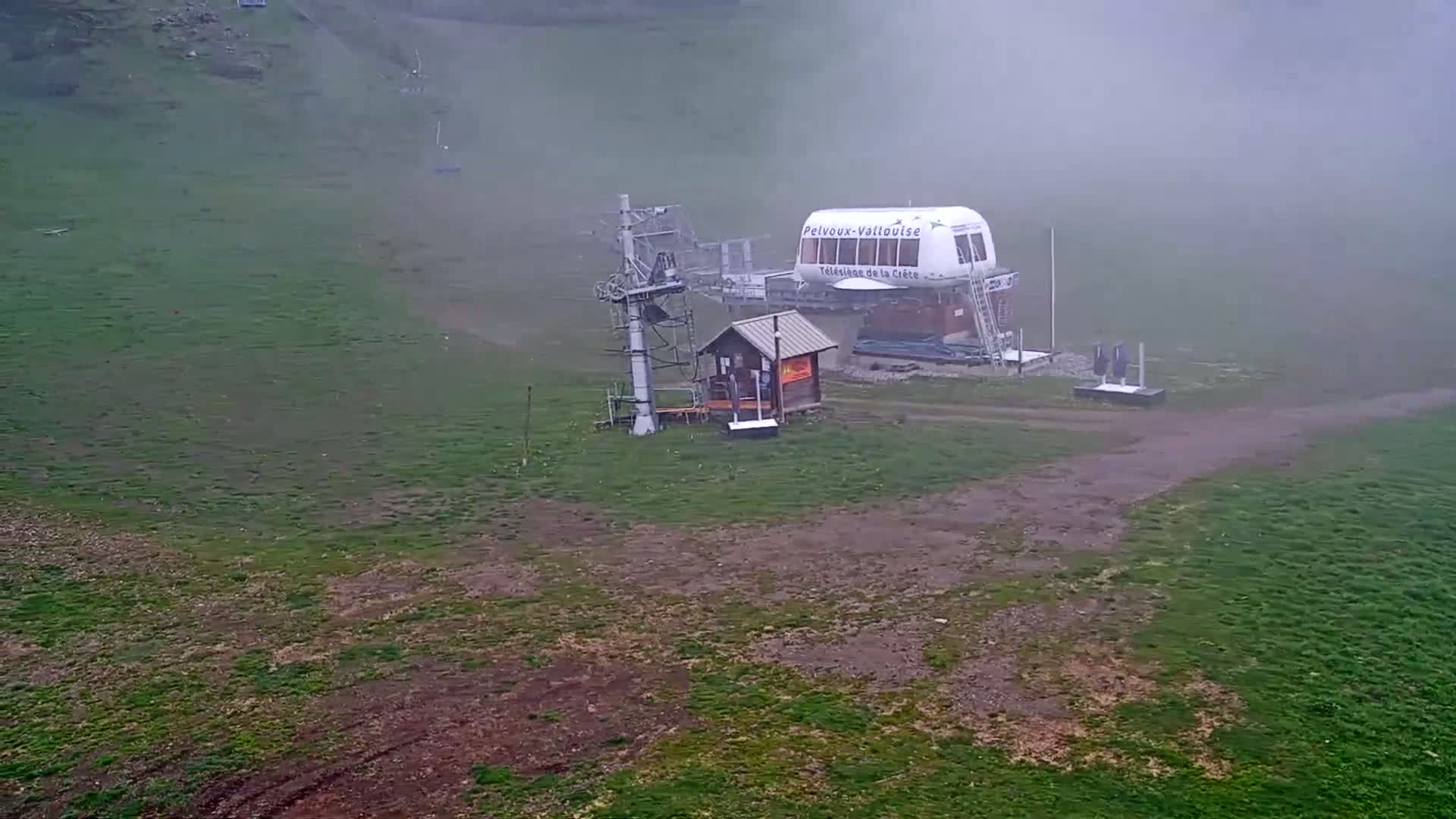 A foggy, grassy hillside features a ski lift station with a small building and a larger gondola car.