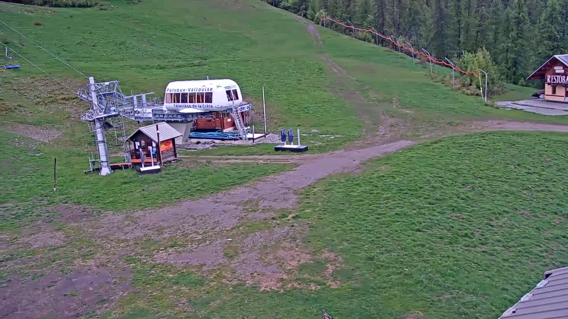 A chairlift base station and small building sit on a grassy hill, with a dirt path leading to a nearby structure under an overcast sky.