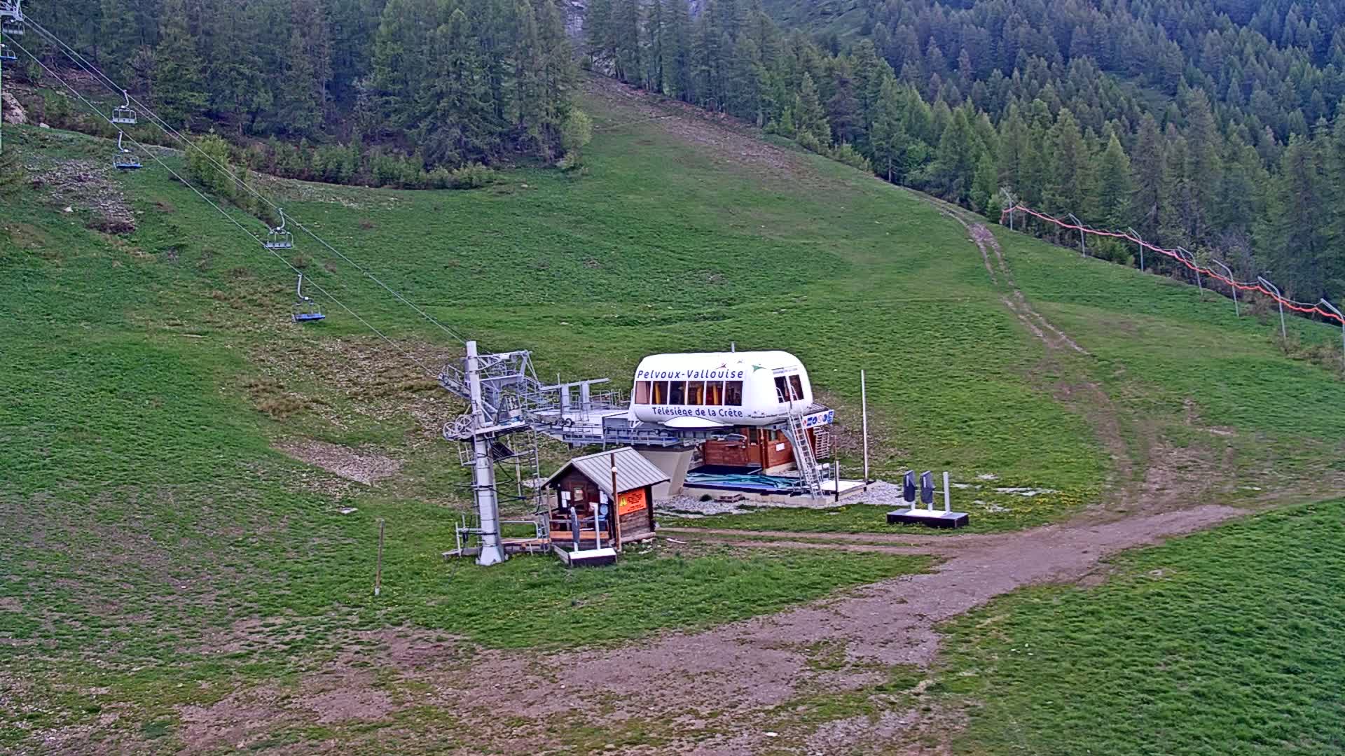 A chairlift station with several empty chairs sits on a grassy hillside amongst a coniferous forest under overcast skies.
