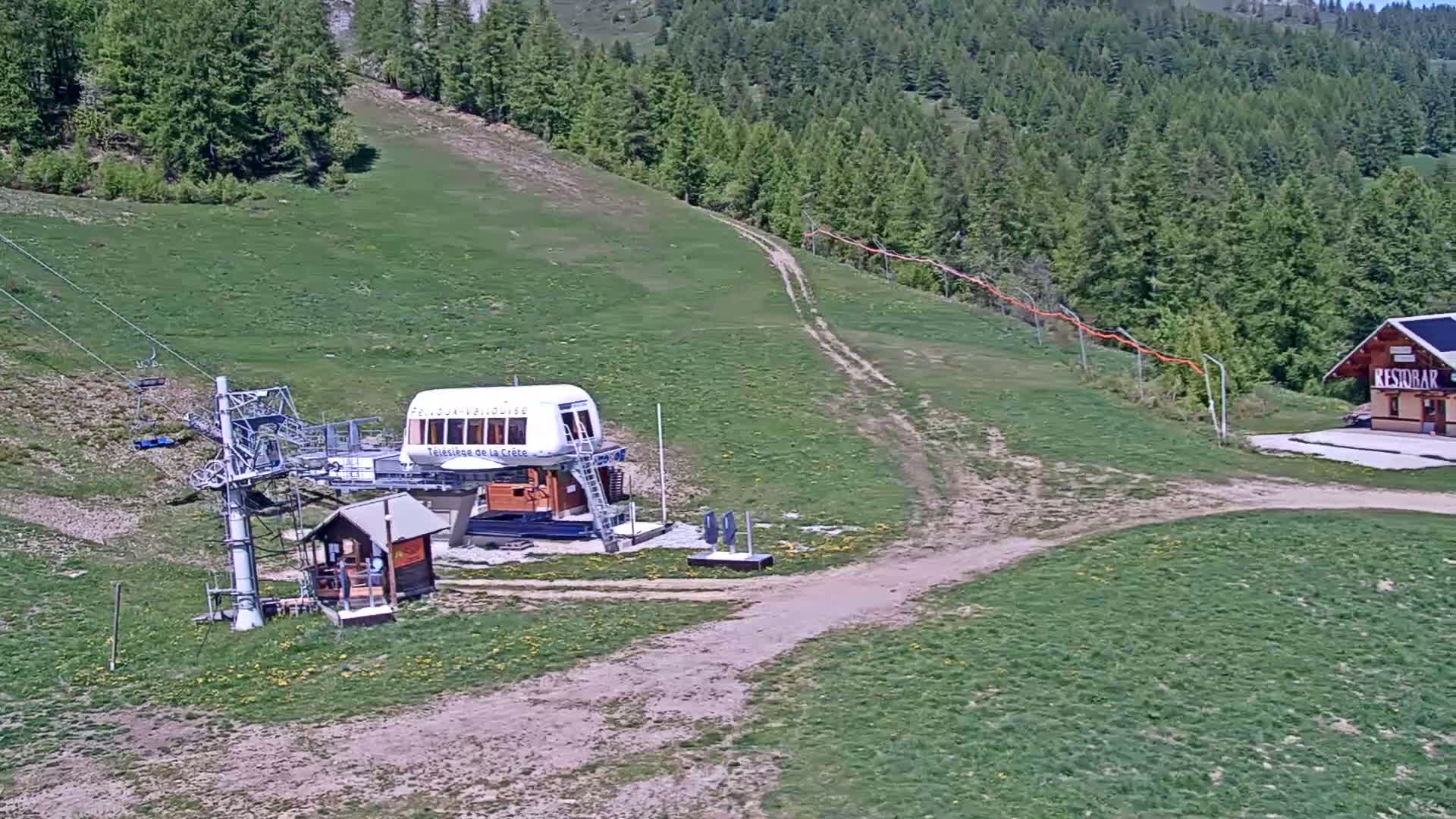 A chairlift base station with a small building sits on a grassy hillside below a forest, under sunny conditions.