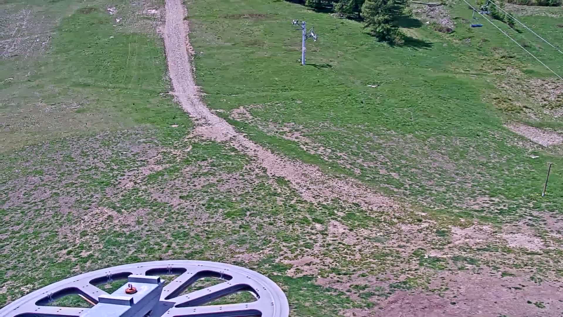 A large wheel is in the foreground, overlooking a grassy hillside with a dirt path and a ski lift in the distance on a sunny day.