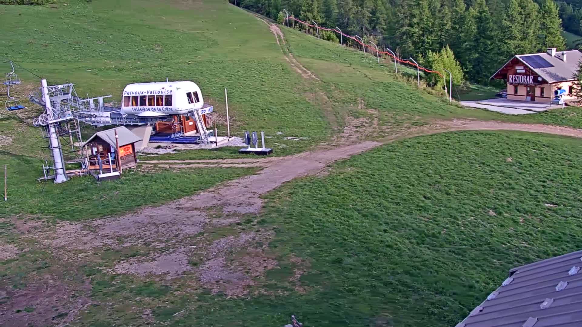 A chairlift station, a small building, and a grassy hillside are visible on a sunny day.