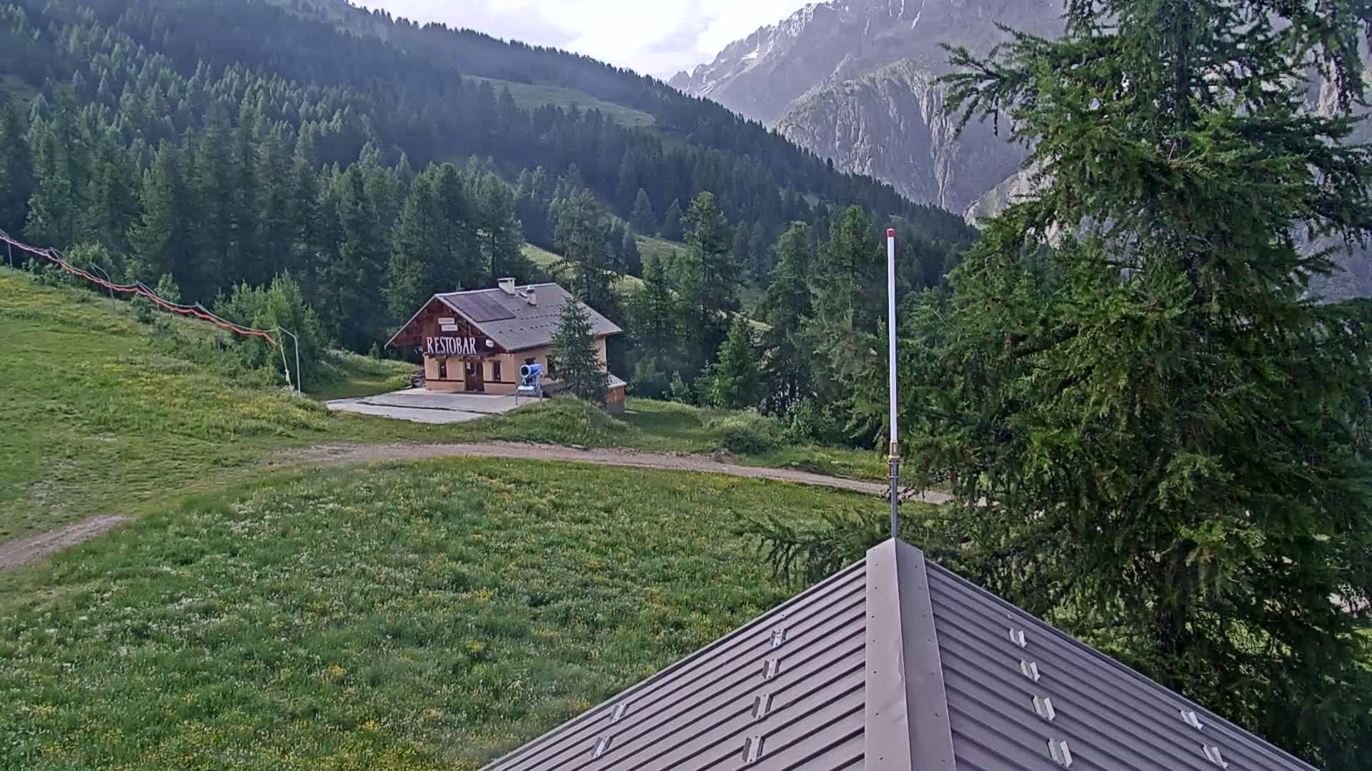 A small building sits on a grassy hillside in front of a mountain range under partly cloudy skies.