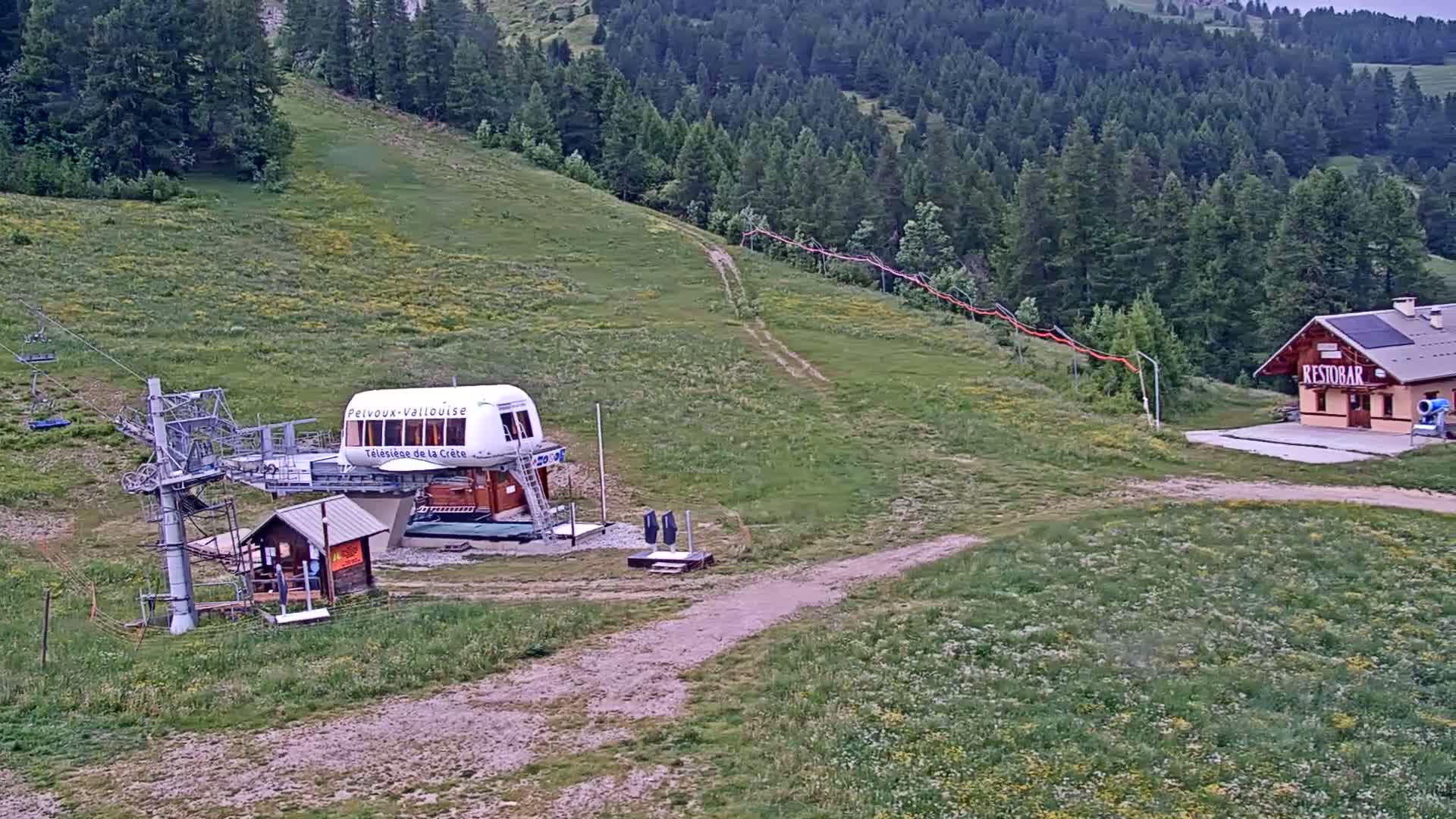 A chairlift station and a small building sit on a grassy hillside overlooking a forest under an overcast sky.