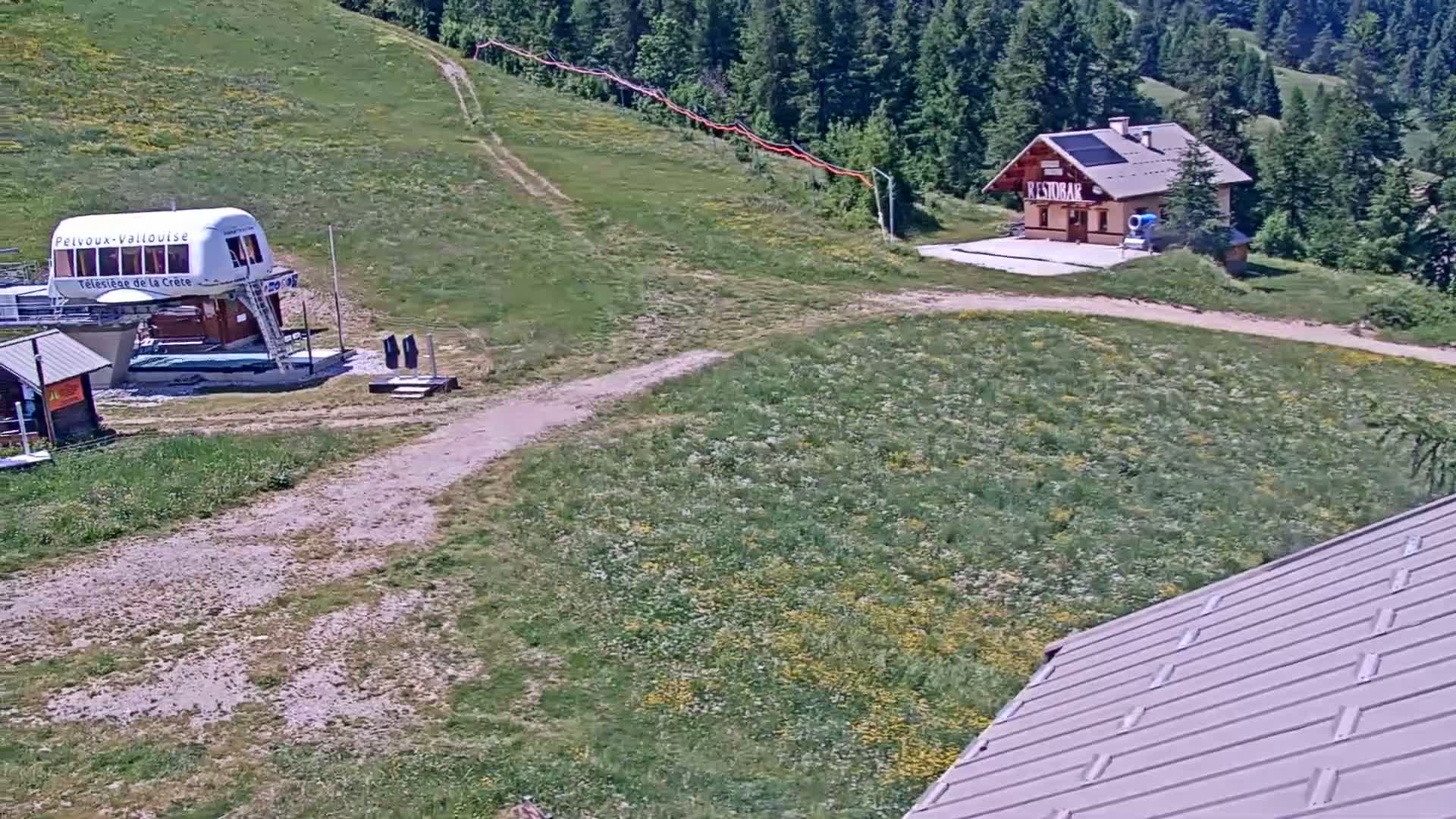 A grassy hillside with a chairlift, small building, and a dirt road is visible under a sunny sky.