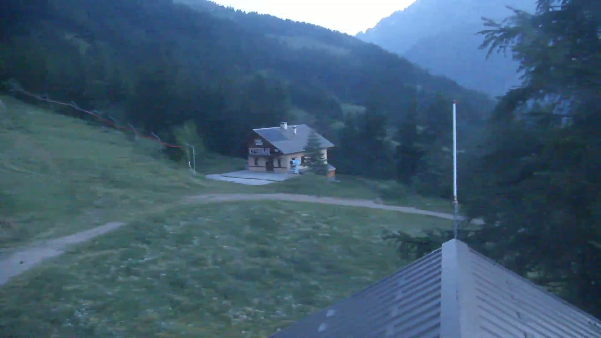 A small house sits on a grassy hillside overlooking a valley in the mountains under a twilight sky.