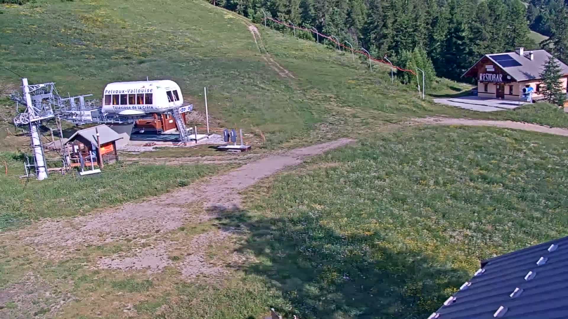 A chairlift station and a small building sit on a grassy hillside under sunny skies, with a dirt road leading between them.