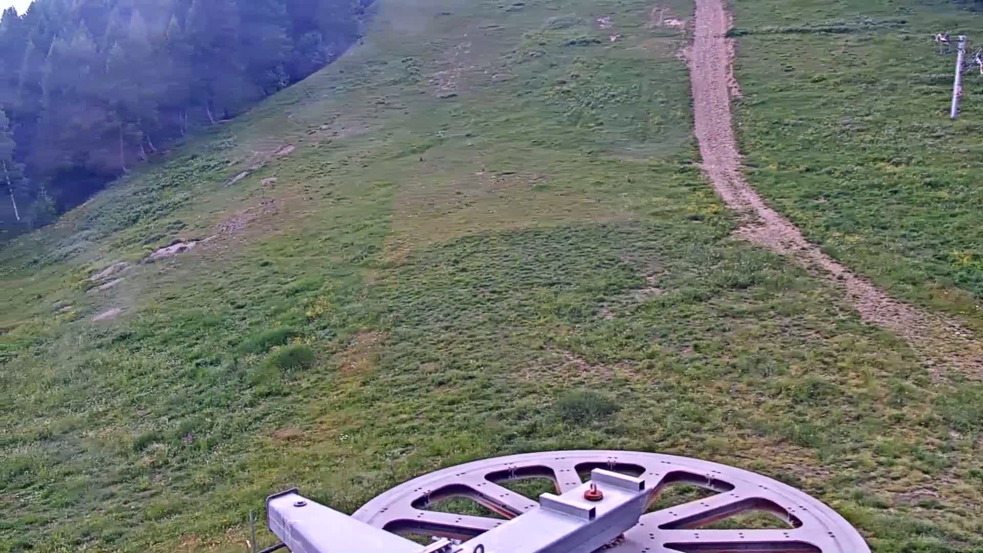A large wheel of a chairlift is visible in the foreground overlooking a grassy hillside with a dirt path leading up to a forest in the background, under mostly cloudy skies.