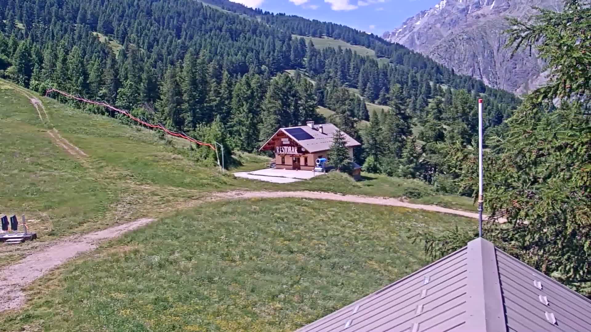 A small building sits on a grassy hillside surrounded by evergreen trees and mountains under a sunny sky.