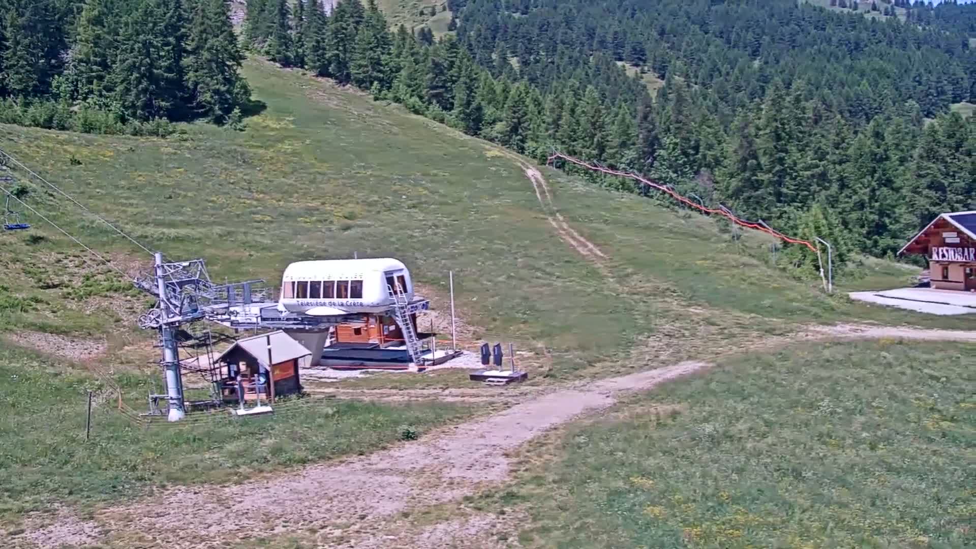 A chairlift station and a small building sit on a grassy hillside overlooking a dense forest on a sunny day.