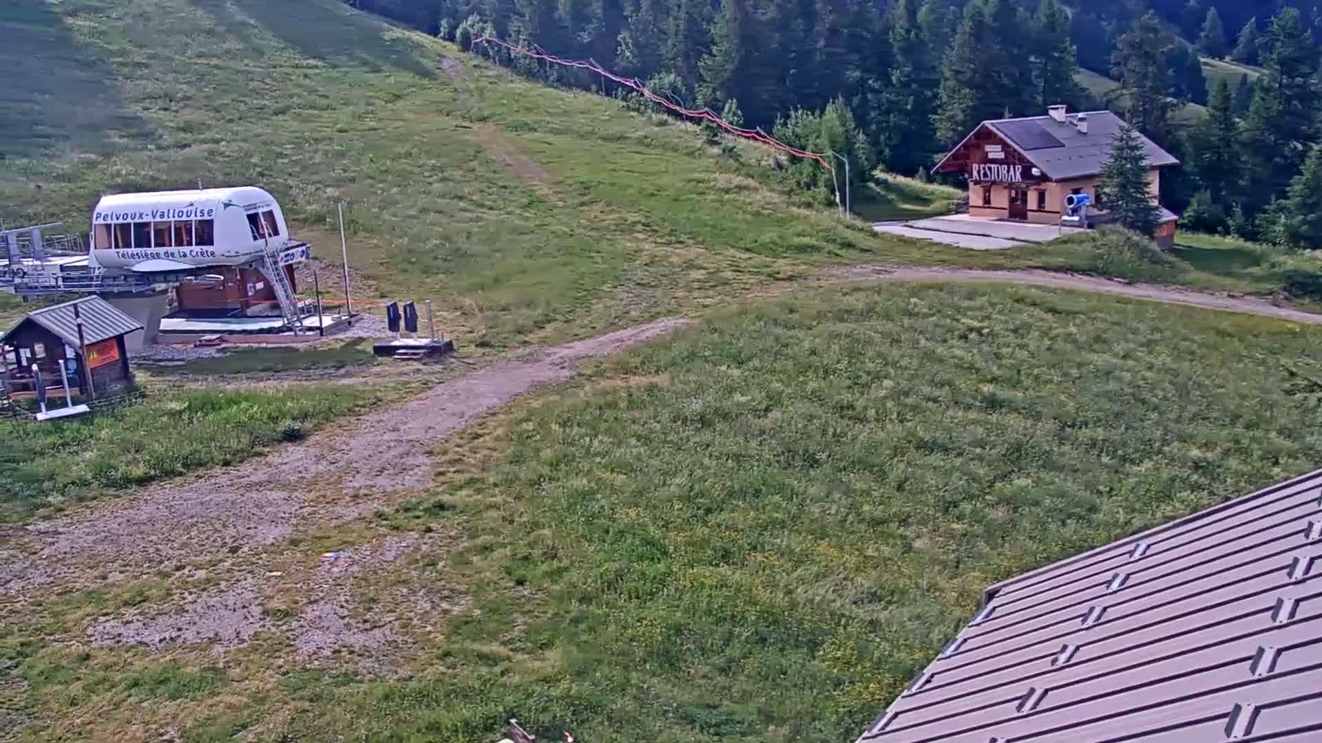 A chairlift base station and small restaurant sit on a grassy hillside under a clear sky, with a portion of a building's roof in the foreground.
