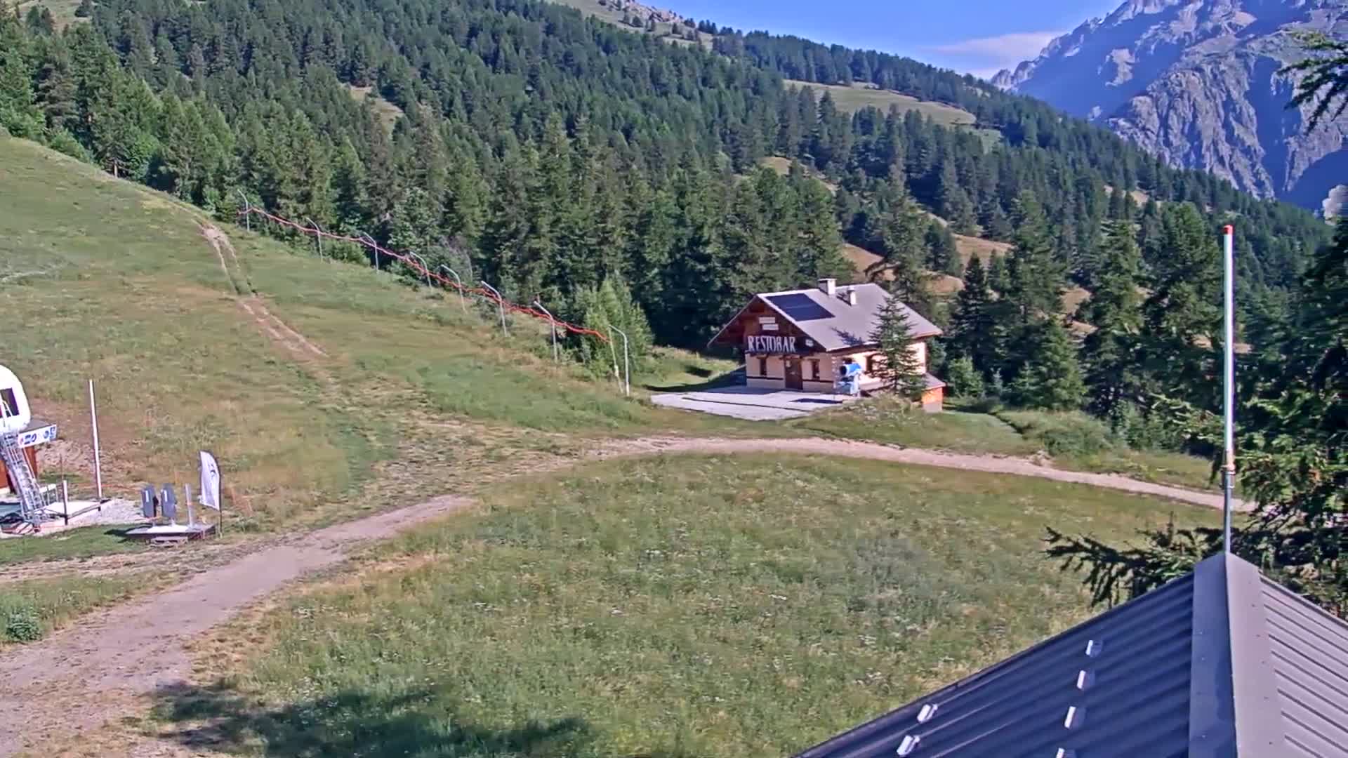 A small building sits on a grassy hillside overlooking a mountain range under sunny skies.