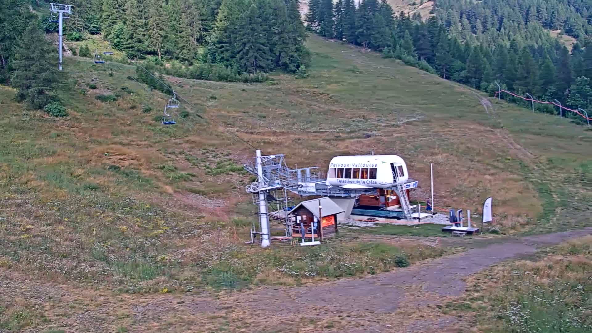 A chairlift station sits on a grassy hillside under a clear sky, with a few empty chairlifts visible on the cable.