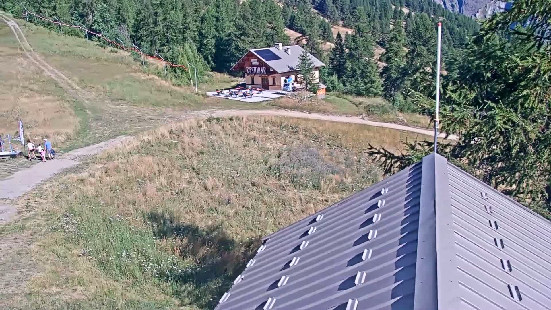 A family walks on a path toward a small building in a sunny, mountainous area with dry, grassy hillsides.
