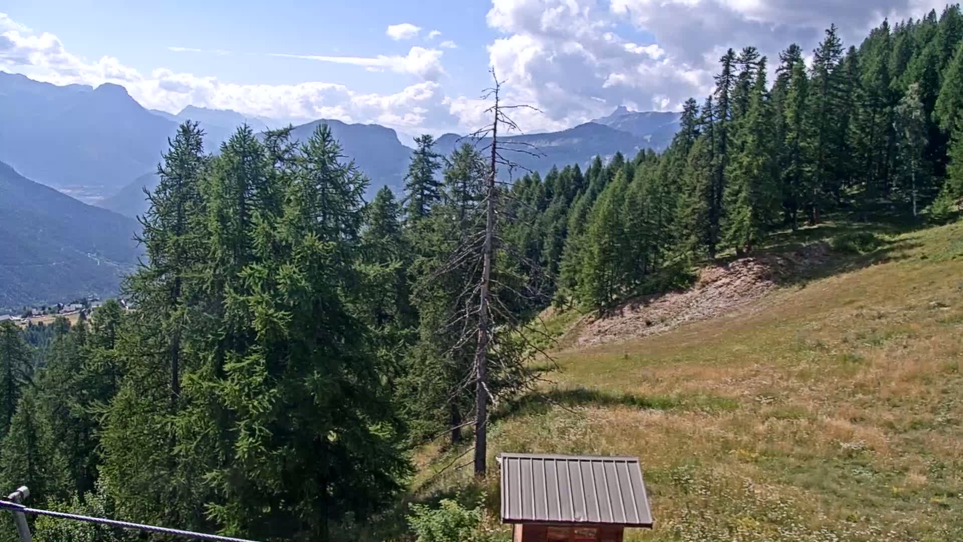 A partly cloudy day reveals a mountainous landscape with evergreen trees, a grassy hillside, and a small shed, overlooking a valley.