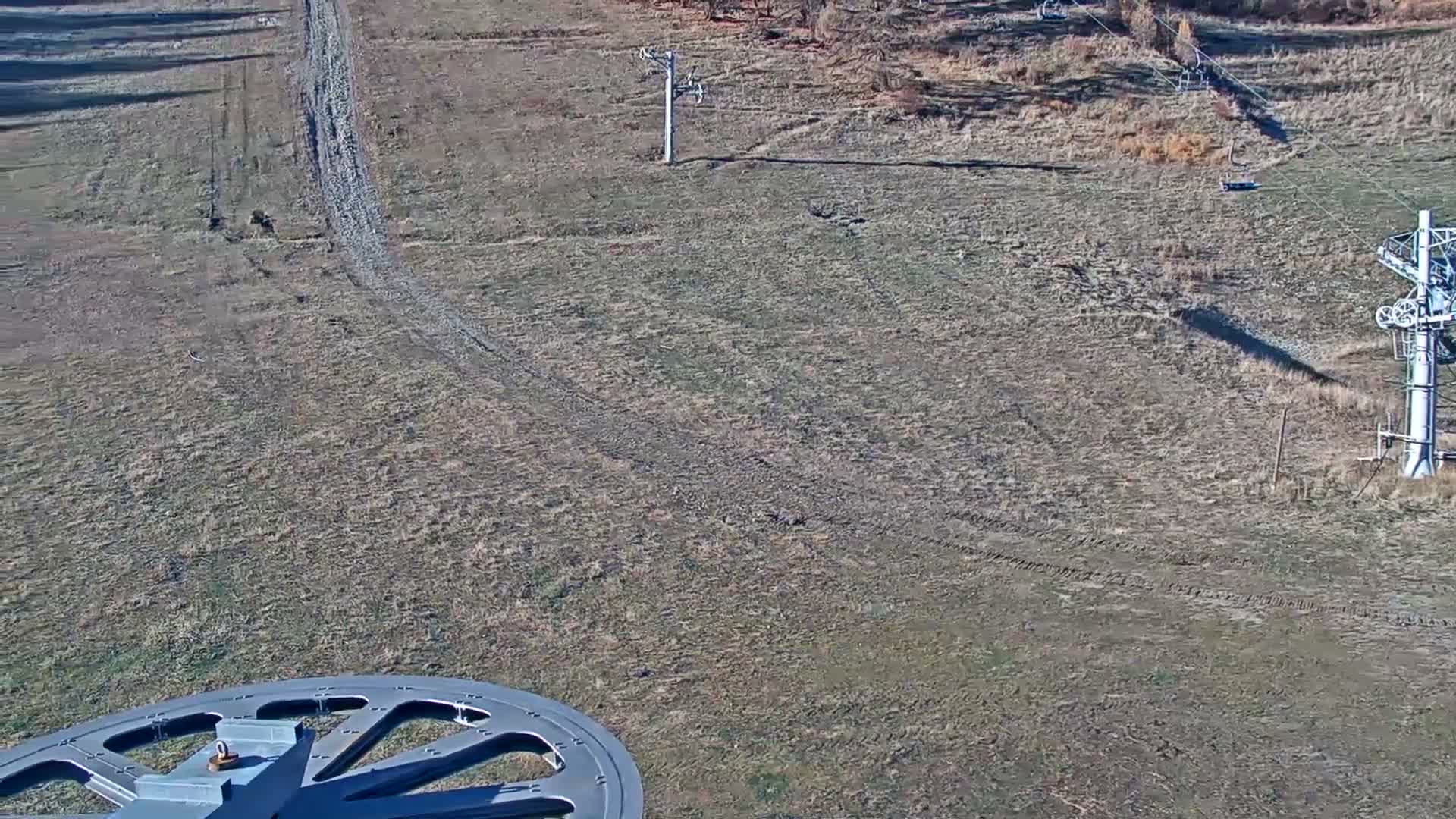 An expansive, dry grassy hillside, featuring several ski lift poles and cables with empty chairs, is bathed in sunlight under a clear sky, with a large gray mechanical structure visible in the lower-left foreground.