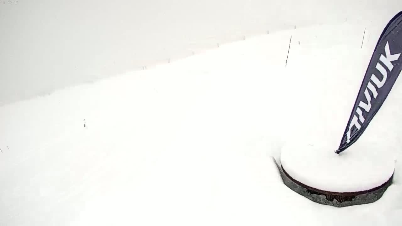 A snow-covered ground with a dark circular object partially buried in the snow, and a dark pennant flag sticking out of it, in a heavy snowstorm.