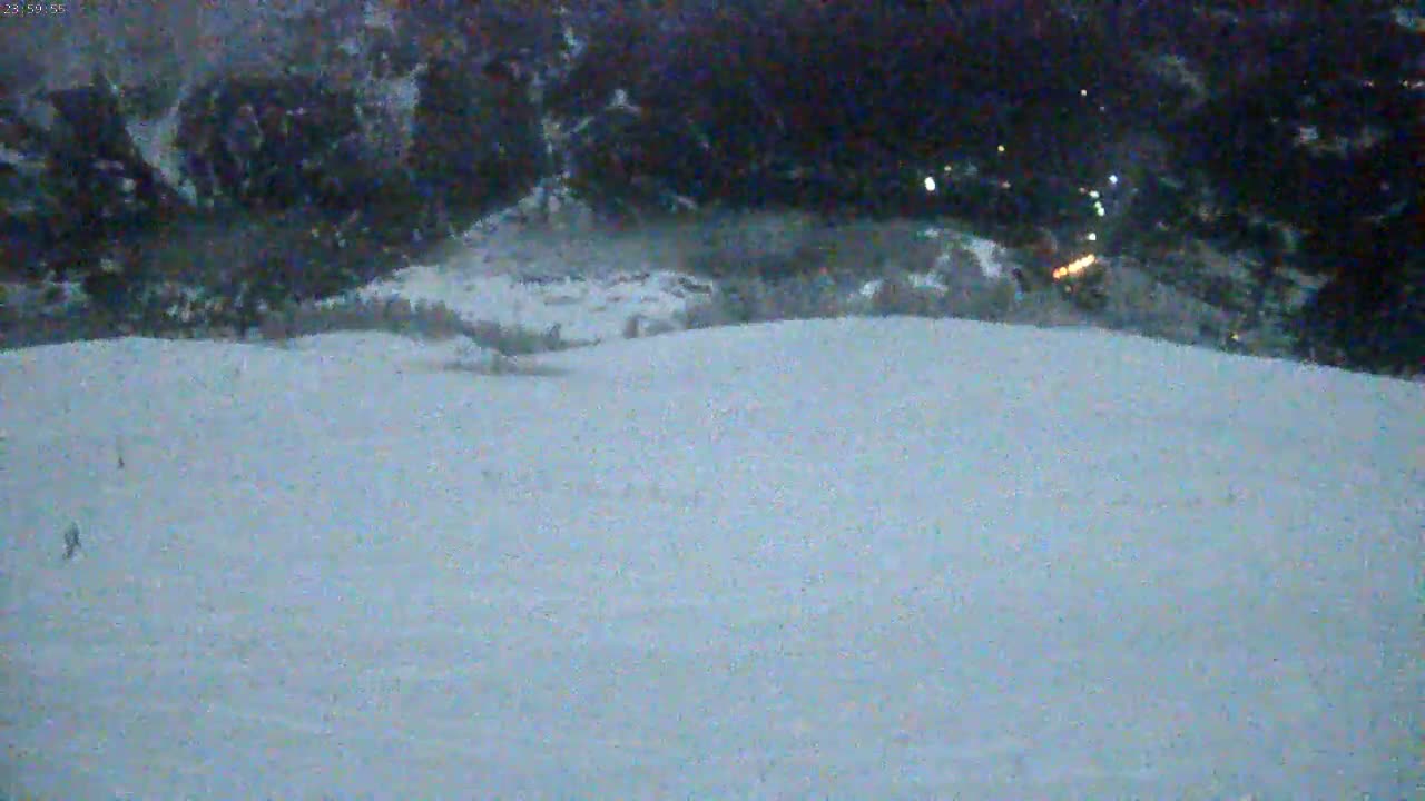 A snow-covered slope at dusk or night, with a few distant lights visible in the background and a couple of small figures in the foreground.