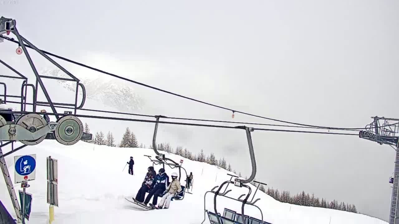 A chairlift ascends a snow-covered mountain in foggy conditions, with skiers visible on the slopes below.