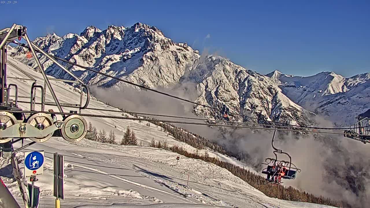 A ski lift carrying skiers ascends a snowy mountain range under a clear blue sky, with low-lying clouds partially obscuring the valley below.