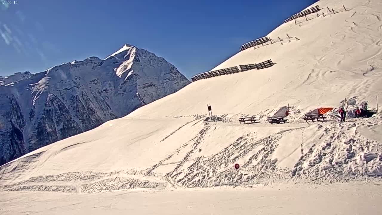 A snow-covered mountain slope with picnic tables and a few people, under a clear blue sky, features a tall snow-capped mountain in the background.